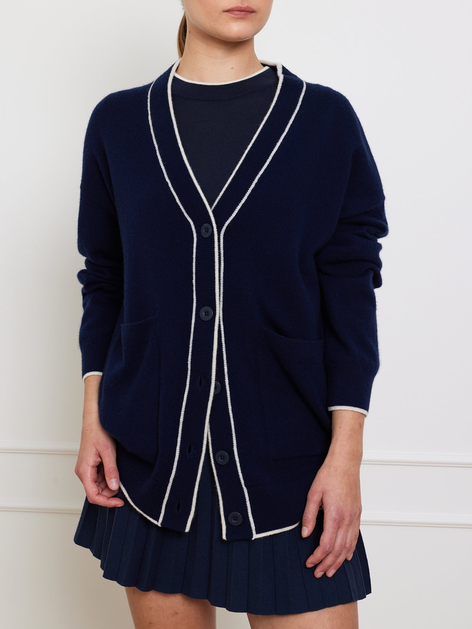 A person elegantly wearing the We Norwegians Club Cardigan in luxurious navy with white trim and black buttons over a dark shirt, paired with a pleated navy skirt, stands against a white wall indoors.