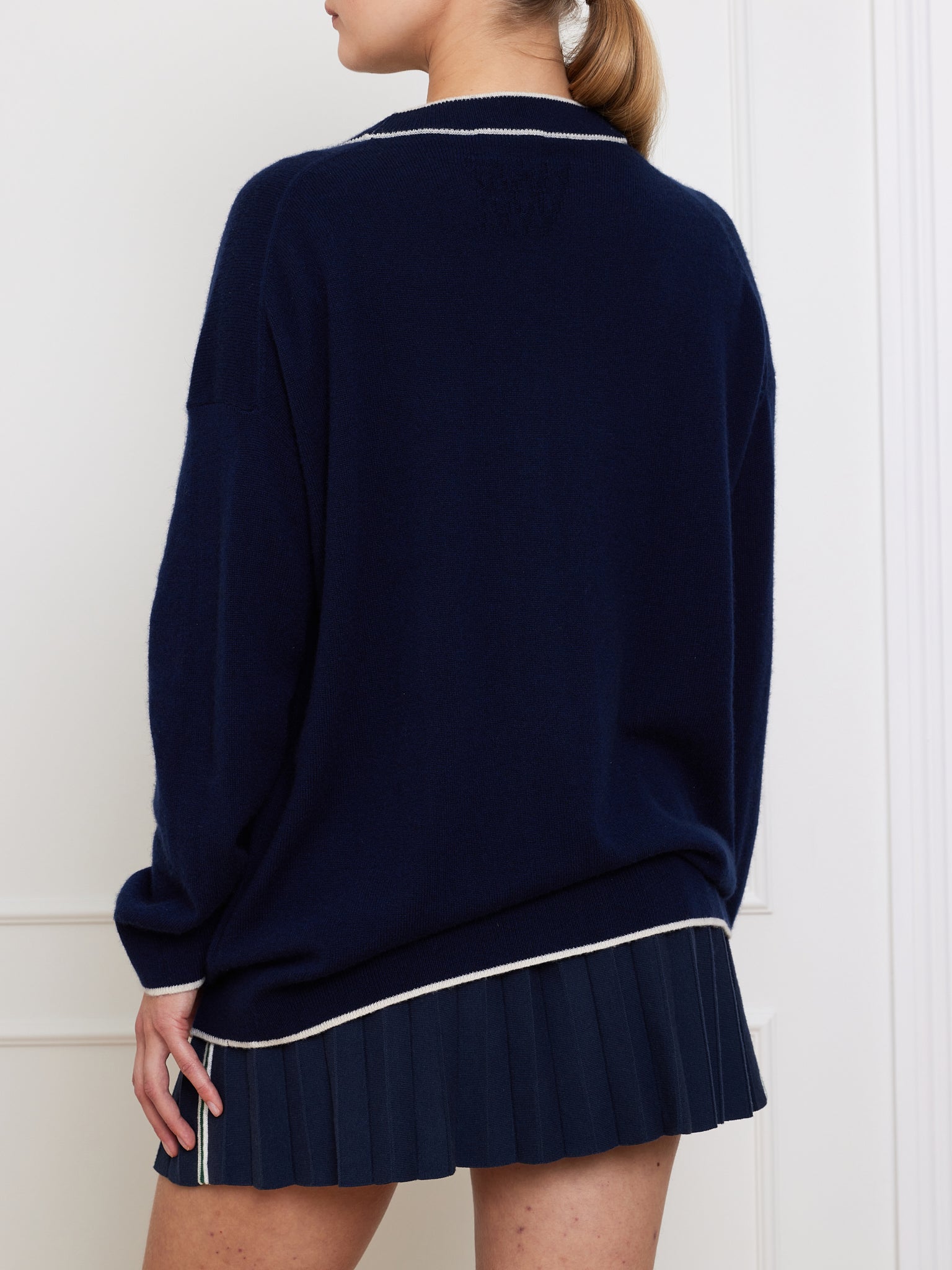 A person in the We Norwegians Club Cardigan, a luxurious navy cashmere piece with white trim, elegantly combines classic style and knitwear. Worn over a navy pleated skirt, their light brown hair is tied back against a pristine white paneled wall.