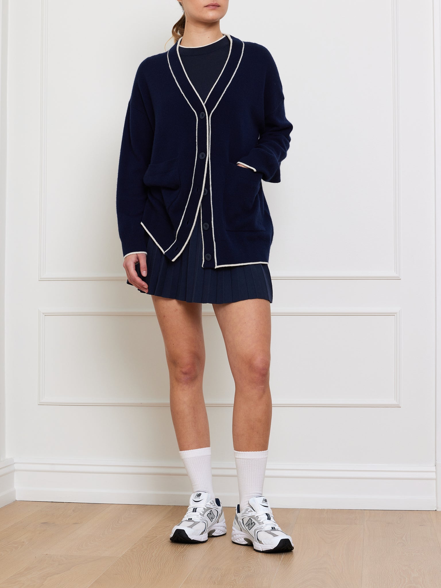 A person wears a We Norwegians Club Cardigan Women in navy with white trim, layered over a dark dress, suggesting luxury knitwear. White socks and sneakers complete the style against white paneled walls and light wood floors. The face of the wearer is not visible.
