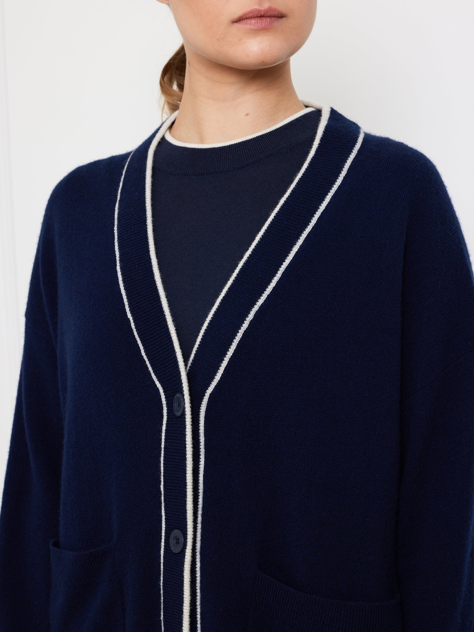 The person is wearing a We Norwegians Club Cardigan in dark blue cashmere with white piping, over a navy shirt. This luxurious knitwear has an oversized fit with front pockets and buttons. Their hair is pulled back against a plain white background.