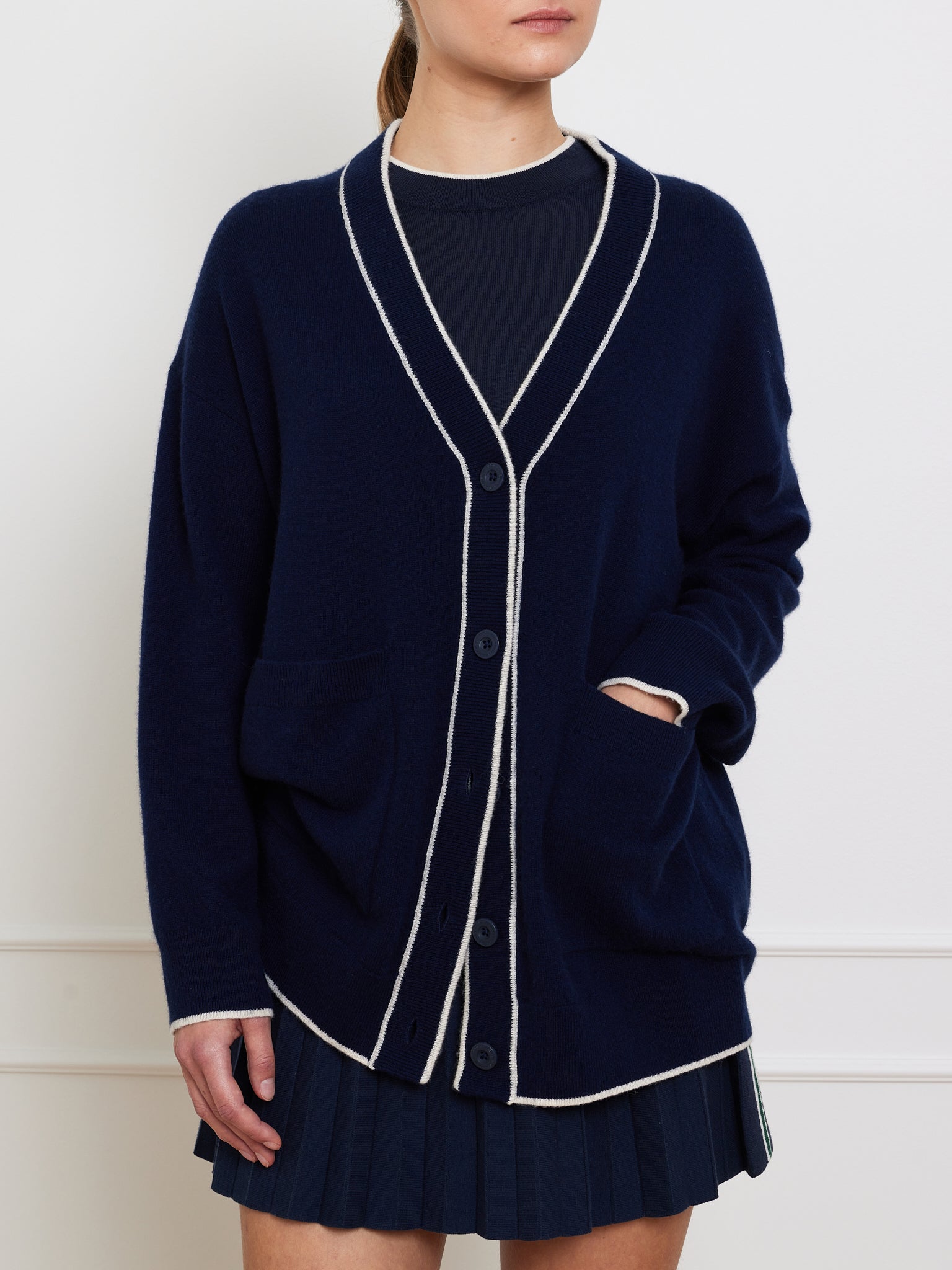 A person is styled in a minimalist and elegant manner, wearing the We Norwegians Club Cardigan Women, an oversized navy blue cashmere piece with white trim, front pockets, and buttons, paired with a pleated skirt.