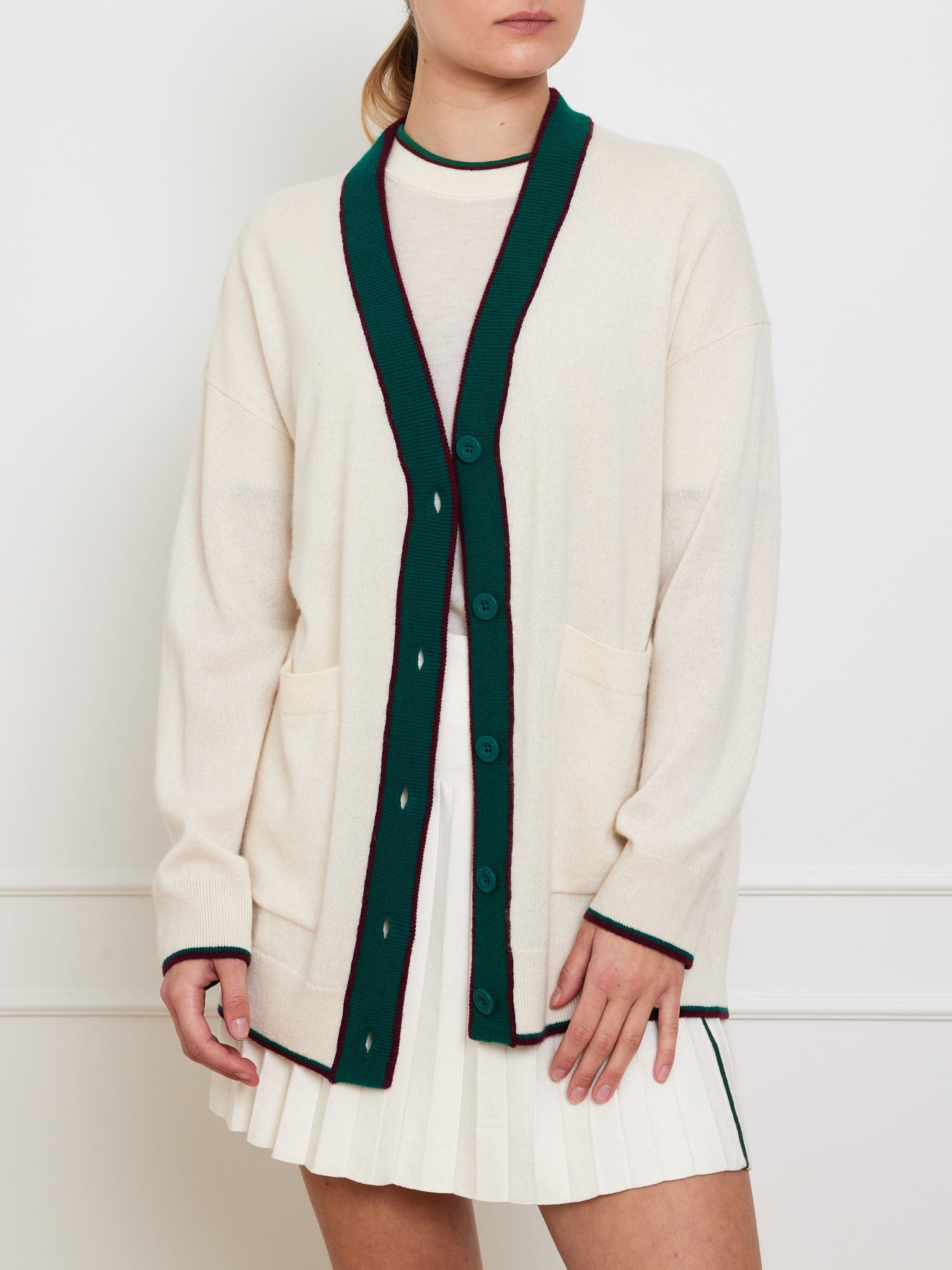 Someone dressed in a We Norwegians Club Cardigan Women, a cream cashmere piece featuring green trim, buttons, pockets, and a deep green border, is paired with a pleated white skirt against a plain background. The person's face is not visible.