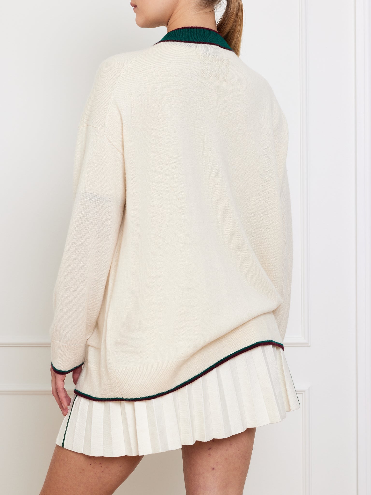 A person showcases a sporty, casual outfit featuring the We Norwegians Club Cardigan Women, a cream oversized sweater with dark trim and pleated skirt. They stand with their back to the camera, highlighting its perfect fit for cooler weather.