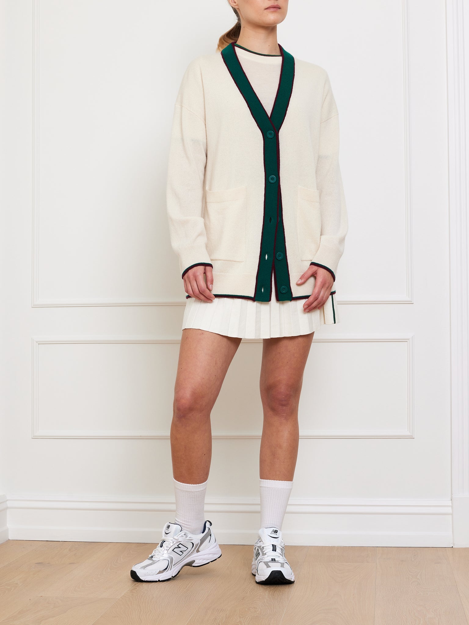 Against a white wall, a person dons the We Norwegians' oversized Club Cardigan Women in cream with black trim, over a pleated white skirt. Completing their sporty yet luxurious look are white sneakers and tall socks. Their face is not visible.