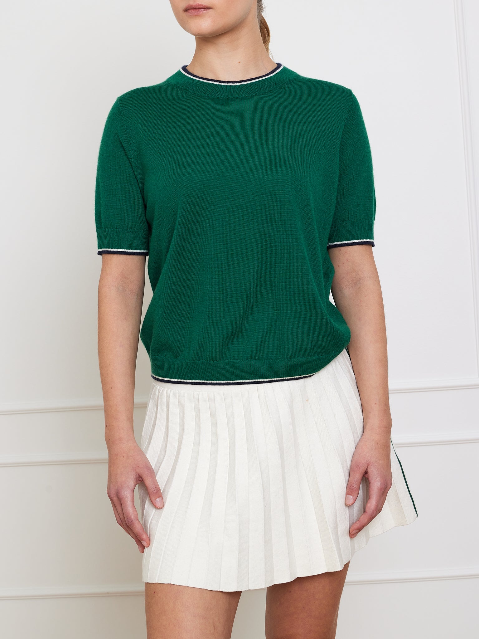 A person models a We Norwegians ensemble—a Club Top Women in green cashmere with white trim paired with a white pleated skirt—against a plain light-colored wall, highlighting the outfit as their arms relax by their sides, concealing their face to emphasize the stylish apparel.