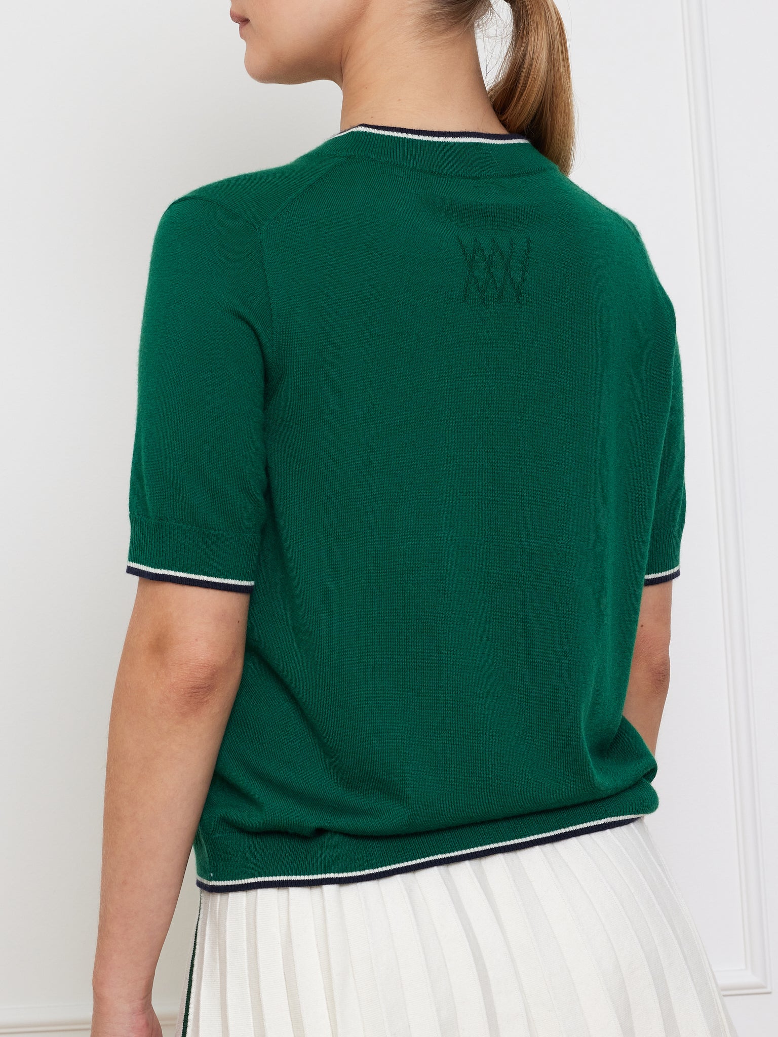 The image features a person with light brown hair in a ponytail, wearing the We Norwegians Club Top Women in green cashmere with white trim and embroidery below the neckline, paired with a white pleated skirt, showcasing relaxed fit designer knitwear effortlessly.