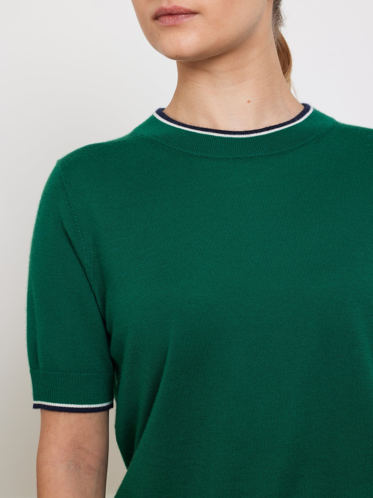 A person is wearing the Club Top Women by We Norwegians, a green short-sleeve cashmere piece featuring black and white trim on the collar and sleeves. Displayed against a plain white background, it highlights the relaxed fit of this designer knitwear.