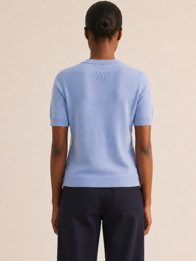 A woman with a neat bun is shown from behind, wearing the We Norwegians Club Top Women - Summer Cashmere in light blue, featuring a stitched "XXX" pattern on the upper back and paired with dark trousers against a plain background.