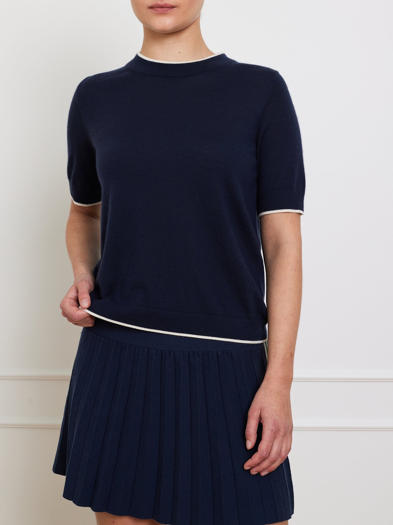 A person is dressed in the We Norwegians Club Top Women, a navy short-sleeve designer knitwear sweater paired with a matching pleated skirt featuring white trim. Set against a white wall, the ensemble offers simple elegance and a relaxed fit perfect for any occasion.