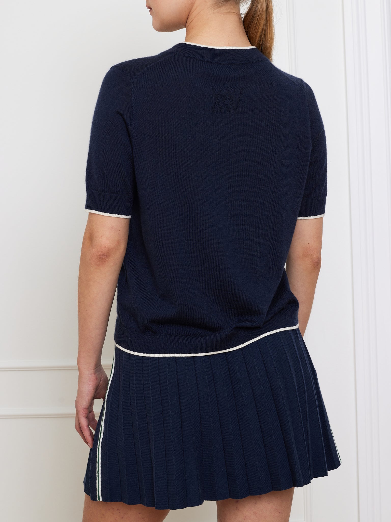 A person indoors against a white wall wears the We Norwegians Club Top Women, a relaxed fit dark blue short-sleeve sweater with white trim, matching pleated skirt, and textured upper back detail reminiscent of designer knitwear.