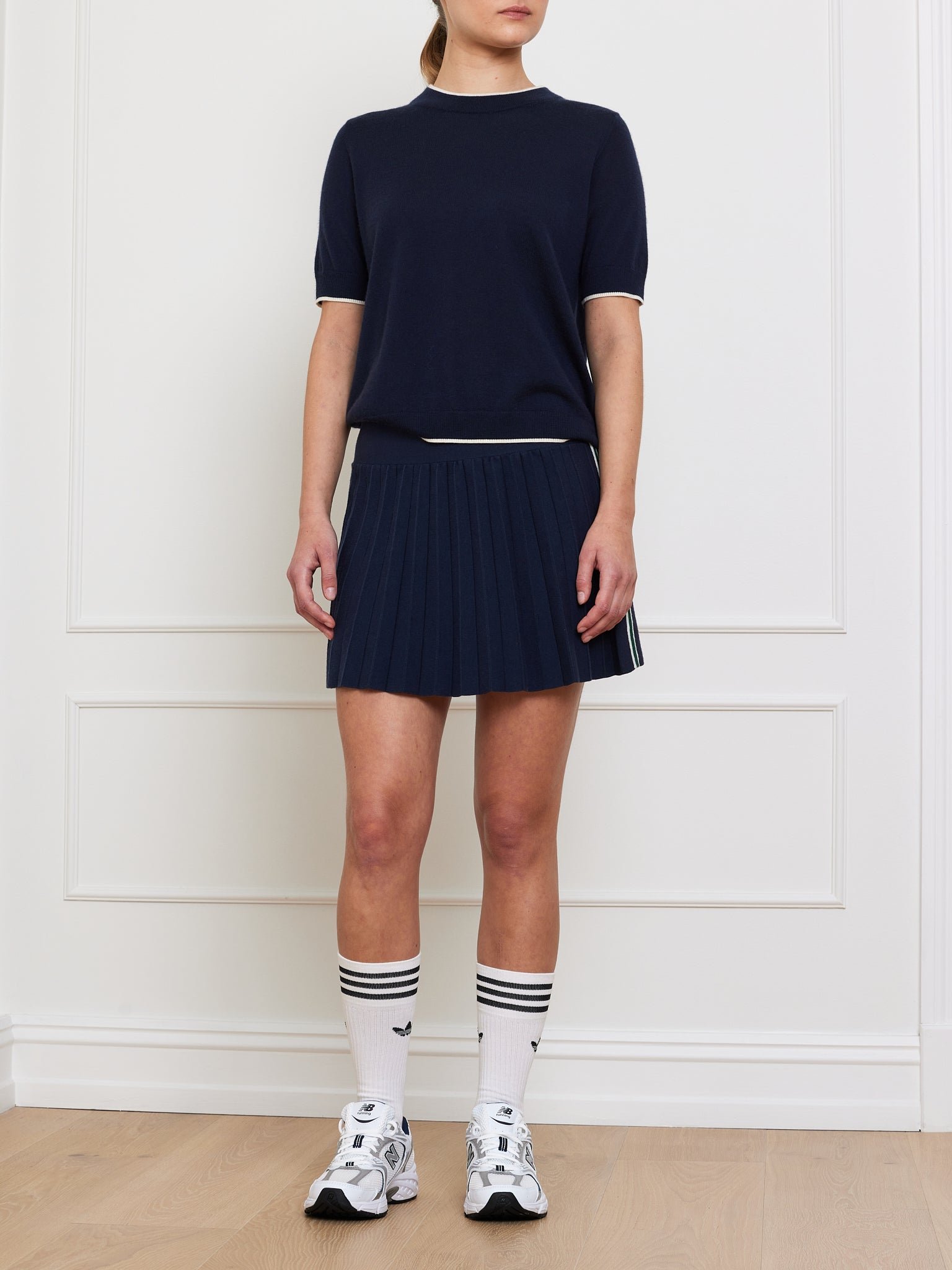 A person stands against a white wall wearing the Club Top Women from We Norwegians, paired with a matching pleated skirt, knee-high white socks with black stripes, and white athletic sneakers. Their face is not visible.