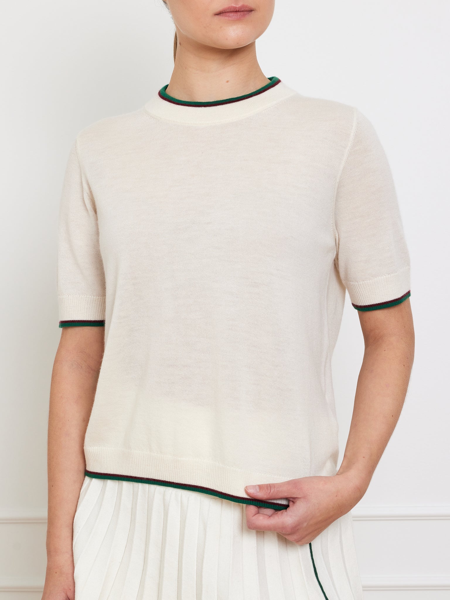 Against a white background, a person wears the Club Top Women by We Norwegians—a cream short-sleeve cashmere top paired with a pleated skirt. This designer knitwear flaunts green and dark accent stripes on the collar and sleeve hems, showcasing relaxed fit and elegance.