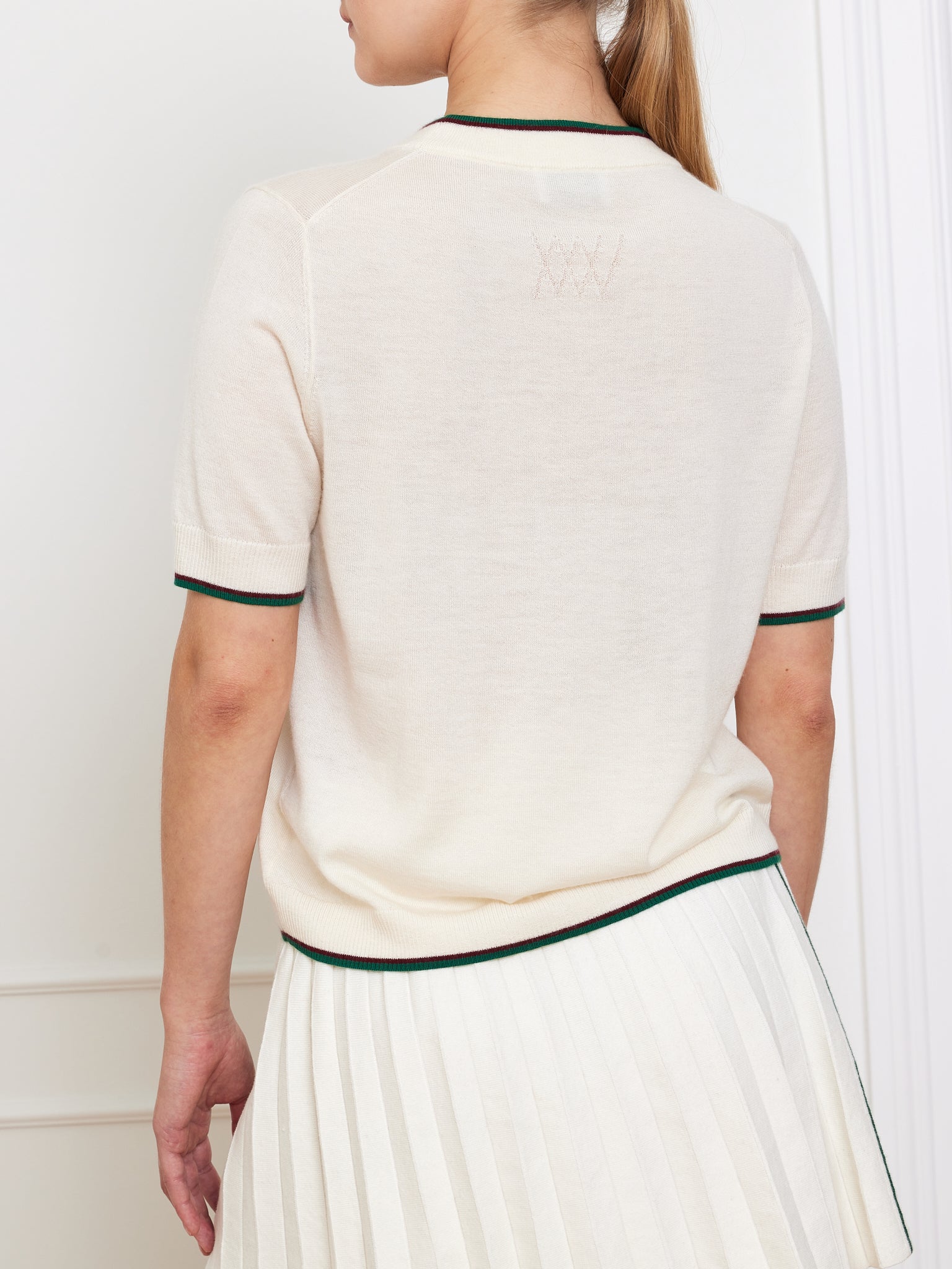 A person with long hair tied back wears the Club Top Women by We Norwegians, featuring a relaxed beige fit with black sleeve and collar trim. They pair it with a pleated white skirt. A partial white wall in the background enhances the serene setting.