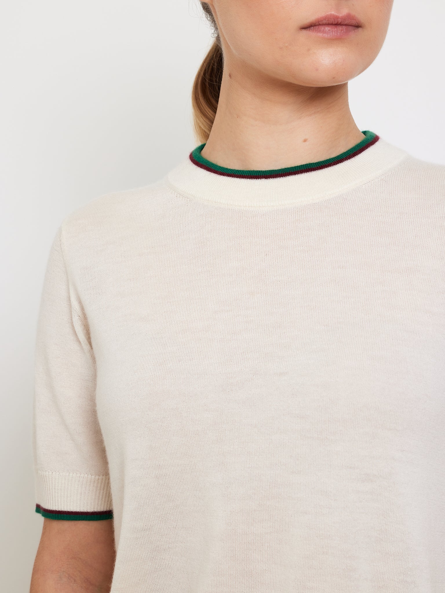 A person with long hair tied back wears the Club Top Women by We Norwegians—a light beige, short-sleeved designer knit sweater with chic green and red trim on the collar and sleeves—set against a plain white background.