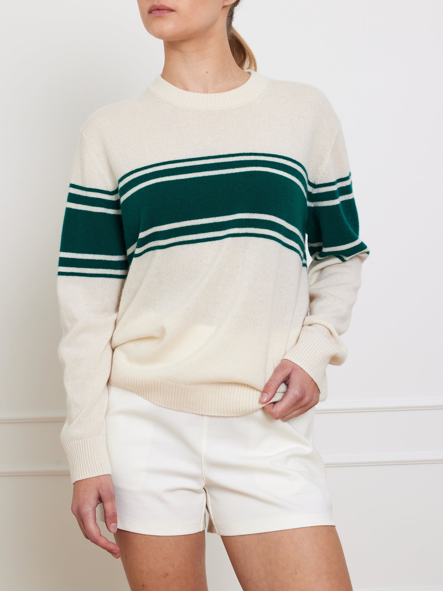 A person models a classic Yacht Sweater Women by We Norwegians, featuring a cream cotton blend with green and white stripes. Paired with white shorts, they stand against a plain white background, one hand resting casually in the sweater's pocket.