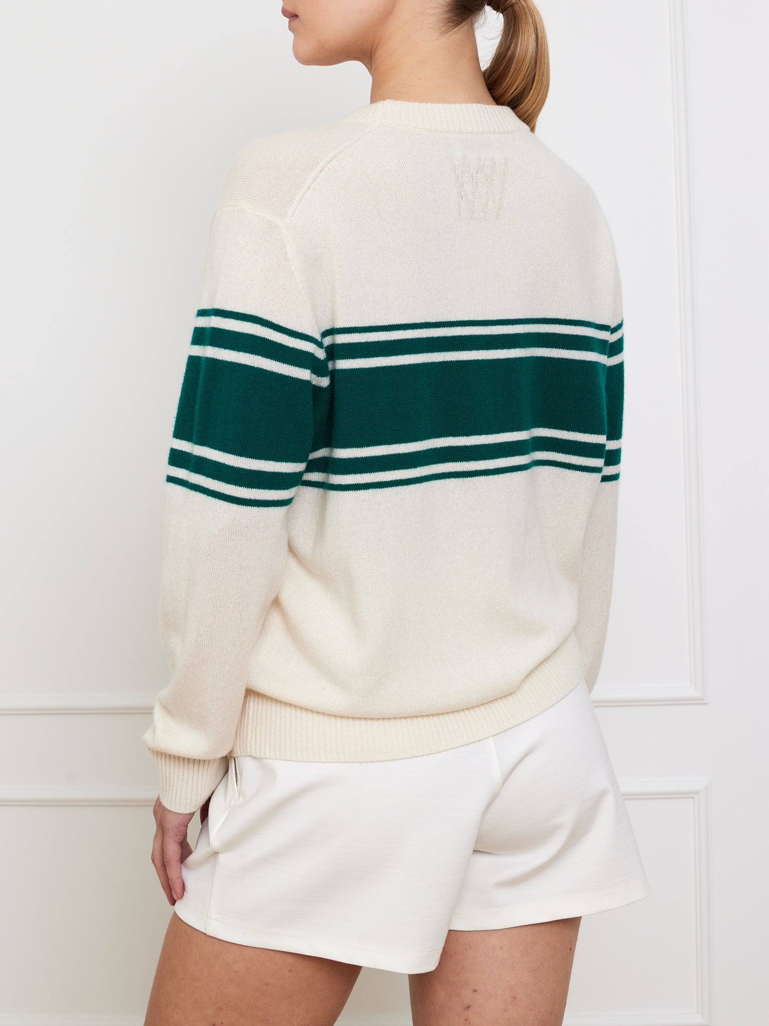 A person with a ponytail wears the We Norwegians Yacht Sweater Women, a classic cream cotton blend with green stripes, paired with white shorts. They stand slightly side-facing against a light-colored, paneled wall.