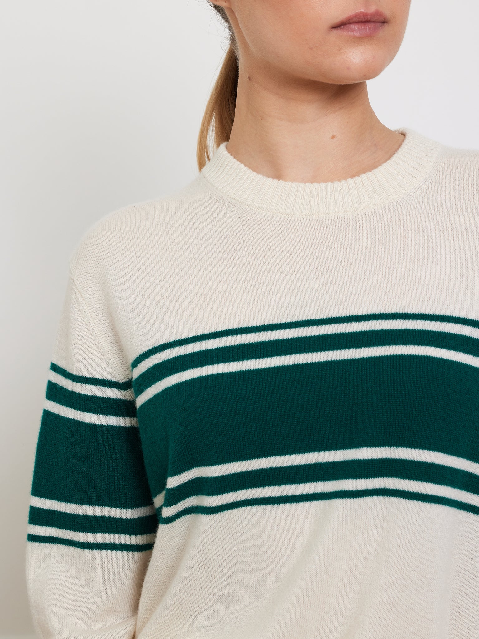 A person wears the Yacht Sweater Women by We Norwegians, a cream-colored cotton-blend classic with green chest stripes and ribbed collar and cuffs. Their hair is tied back, showing only the lower part of their face and upper body.