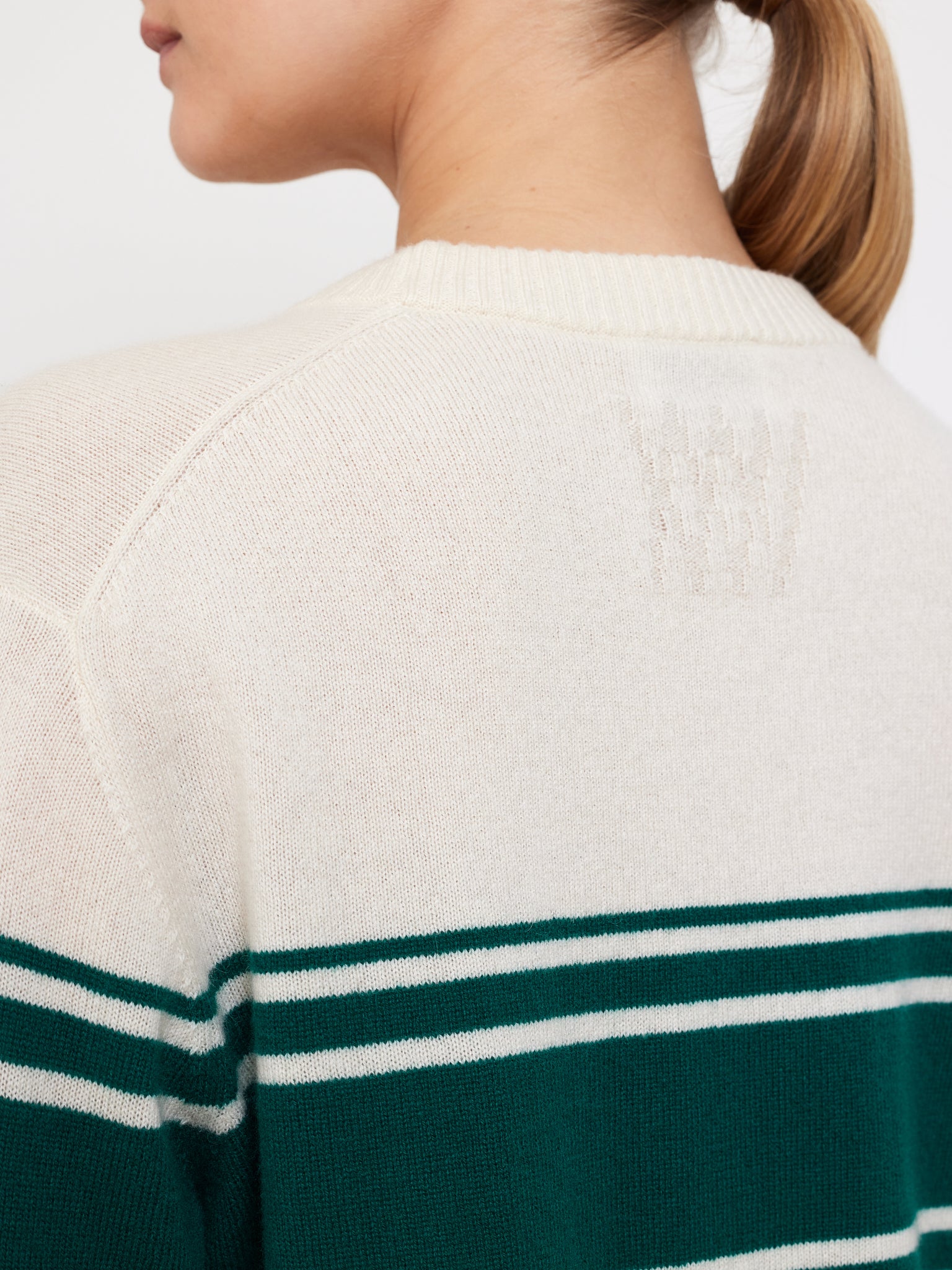A person with light brown hair in a ponytail wears the Yacht Sweater Women by We Norwegians, a cream classic featuring dark green and white horizontal stripes, showcasing its textured pattern from the back.