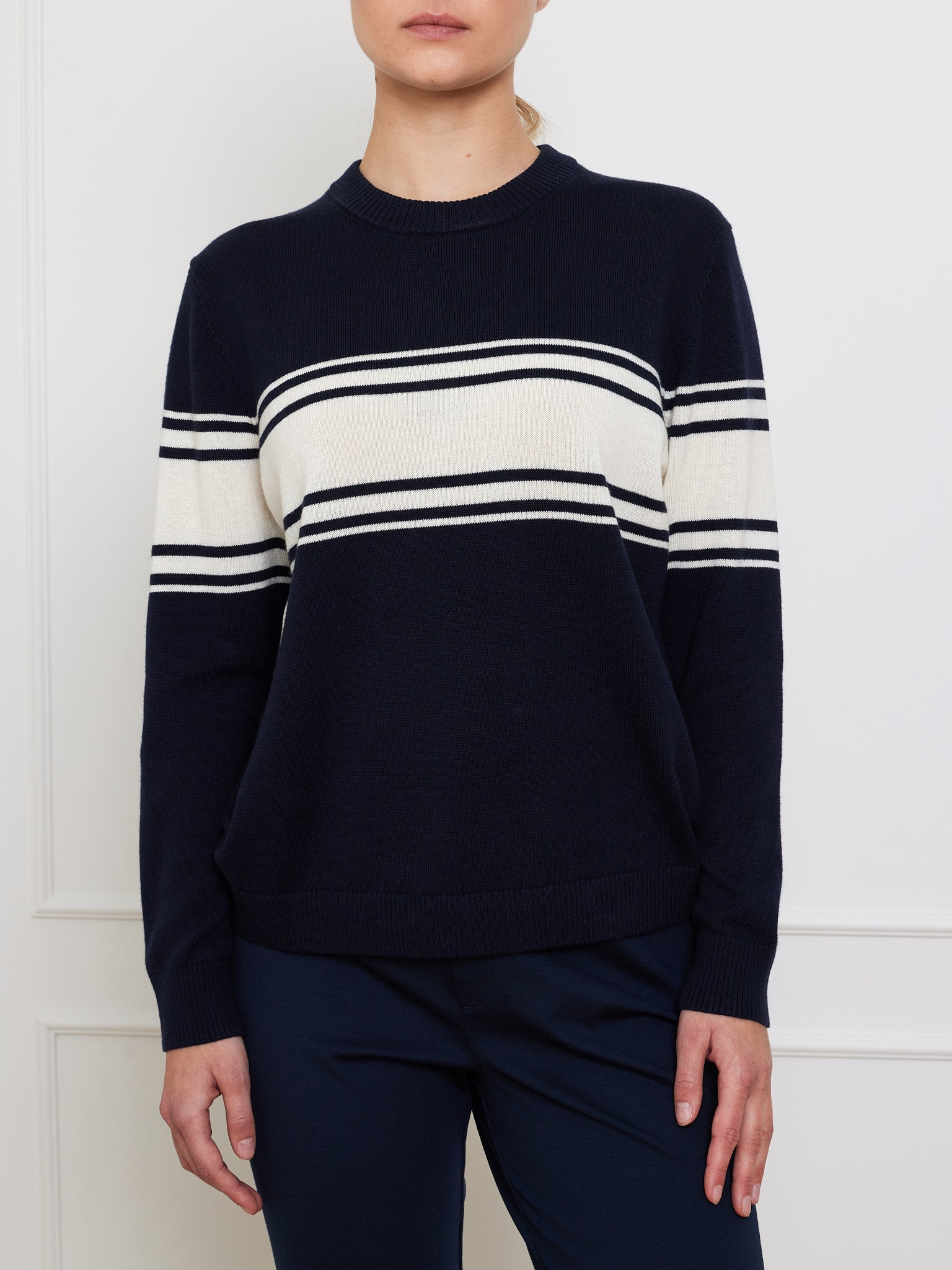 Against a white wall, someone models the We Norwegians' Yacht Sweater Women—a navy cotton blend with white stripes on the chest and arms. The person's face isn't visible, and they wear it with dark blue pants.