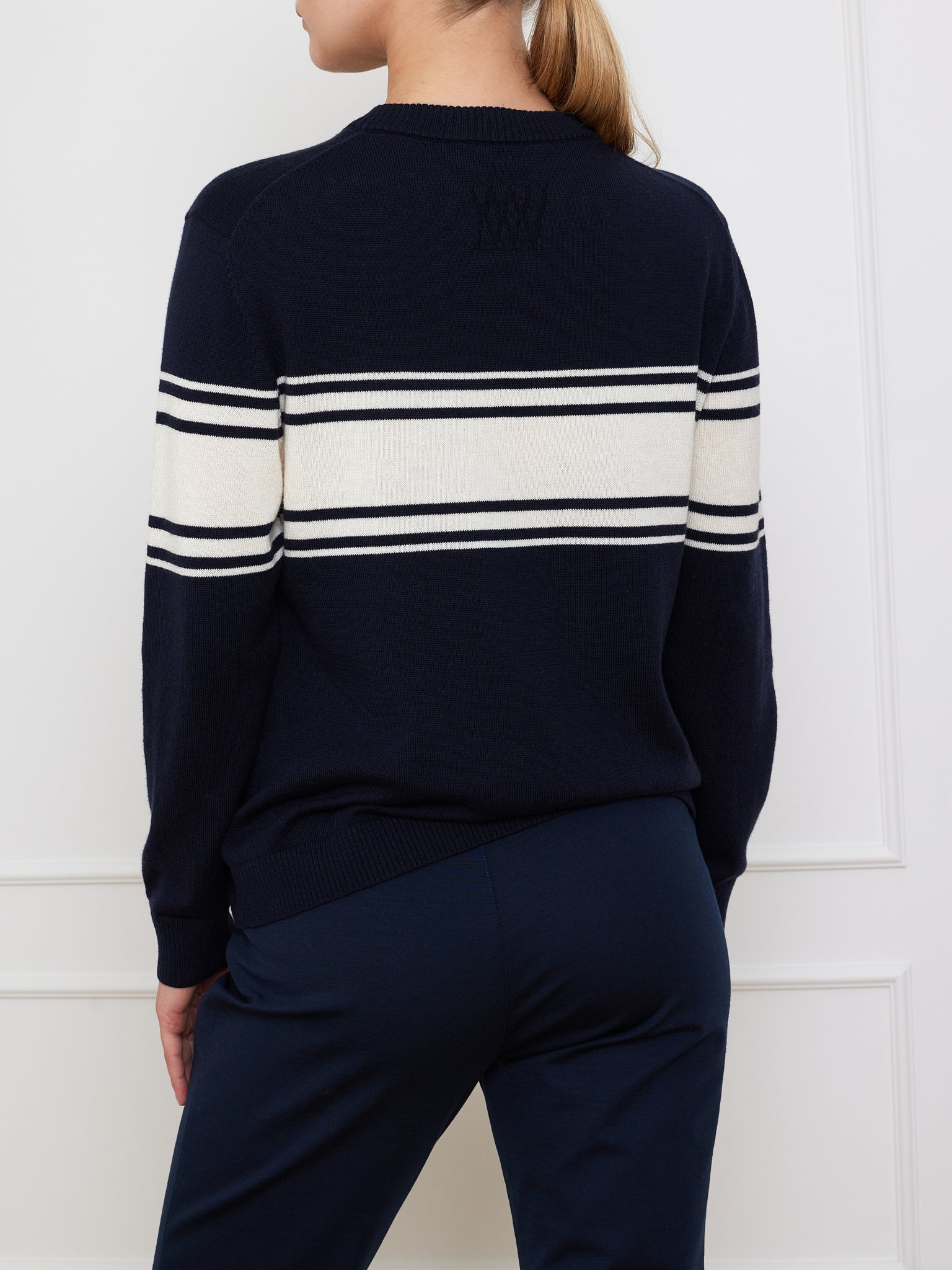 A person with long hair tied back wears the Yacht Sweater Women by We Norwegians, featuring navy blue cotton blend with white and black stripes across the back. They're paired with navy blue pants, standing before a white paneled wall.