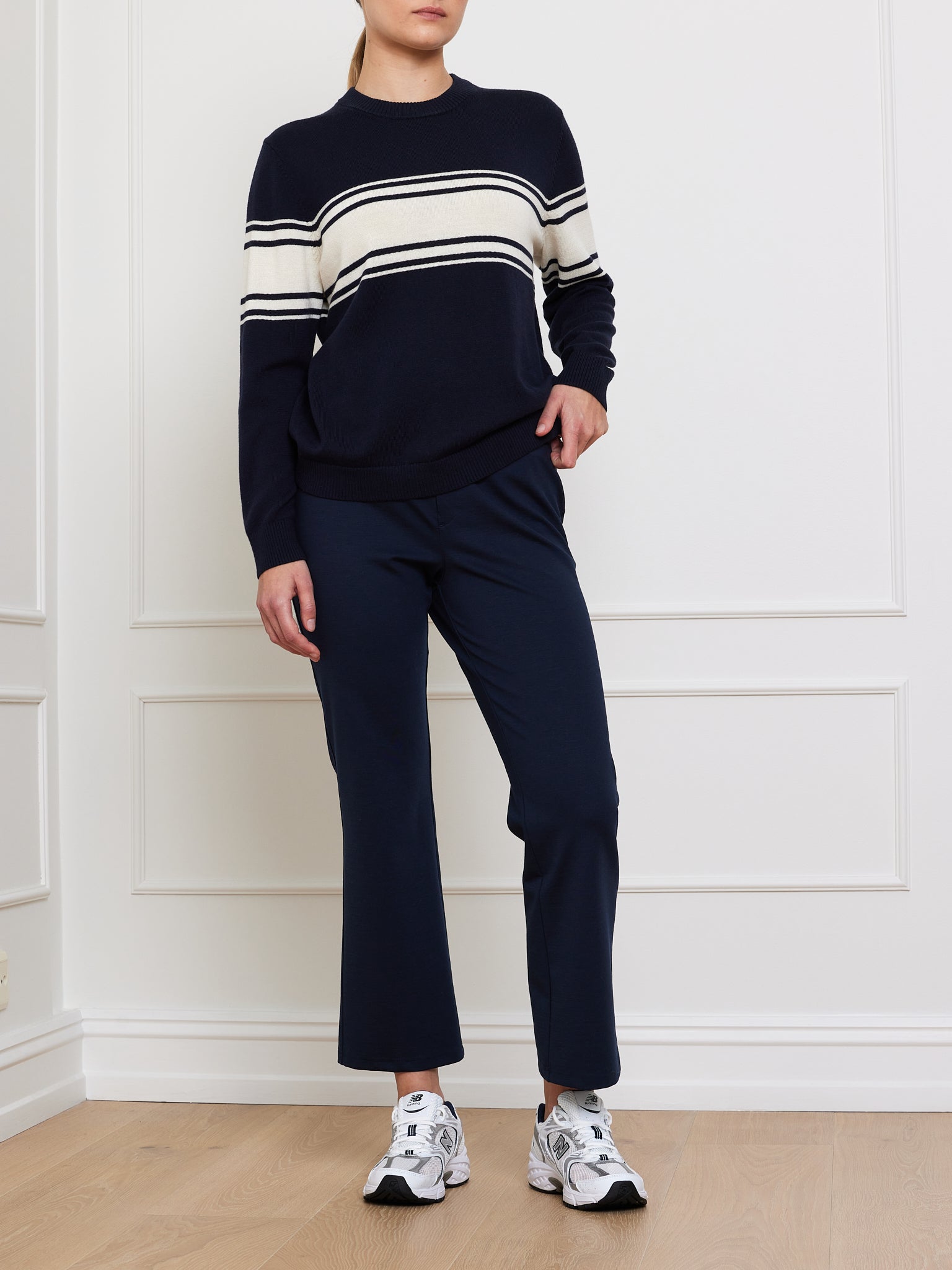A person stands against a white wall, wearing the Yacht Sweater Women by We Norwegians, featuring navy cotton blend with white and beige stripes. They're also in navy pants and white sneakers, but their face is unseen.