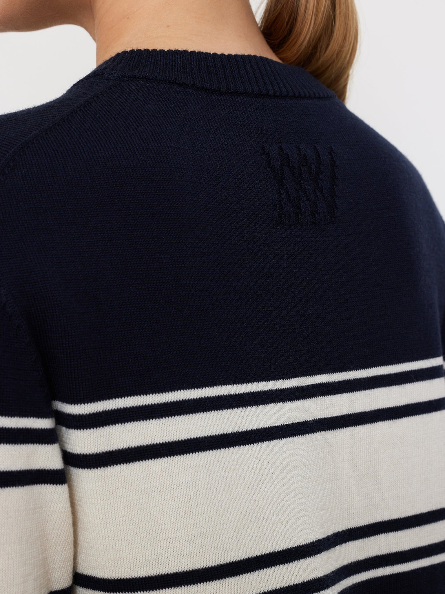 The image shows the back of a person wearing the We Norwegians Yacht Sweater Women, featuring white horizontal stripes on navy merino wool and a textured logo on the upper back. The individual's hair is in a ponytail, highlighting this timeless piece.