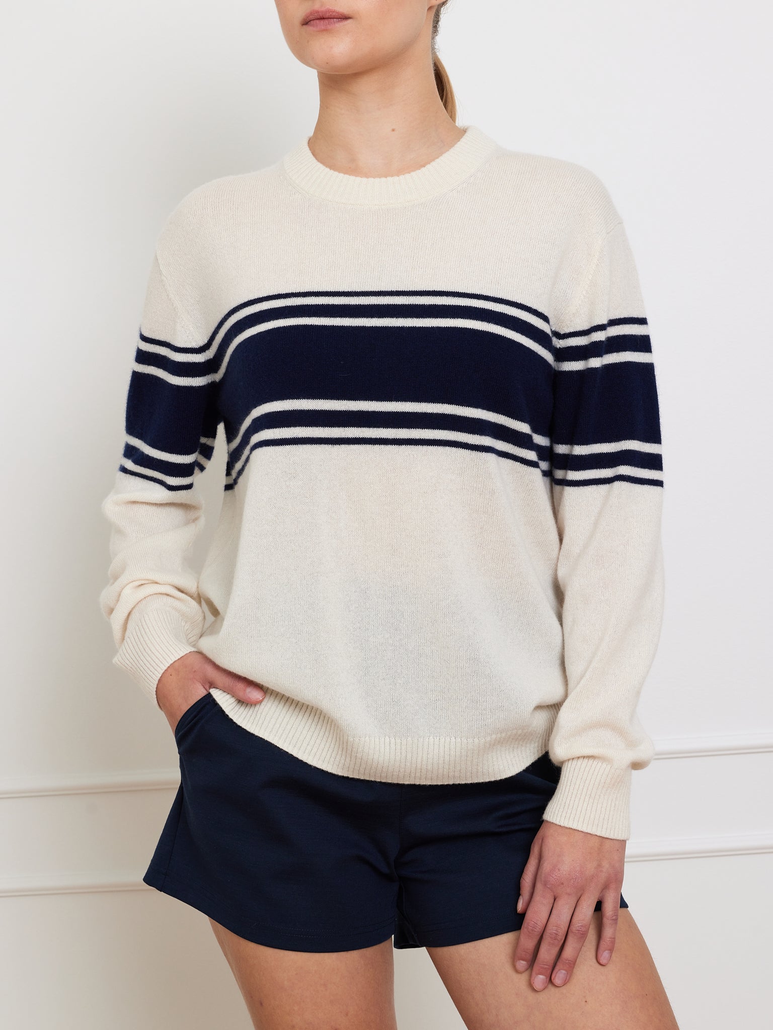 Wearing the Yacht Sweater Women by We Norwegians, featuring cream merino wool with navy stripes, paired with navy shorts. They stand in front of a white wall, hands casually tucked into their pockets.