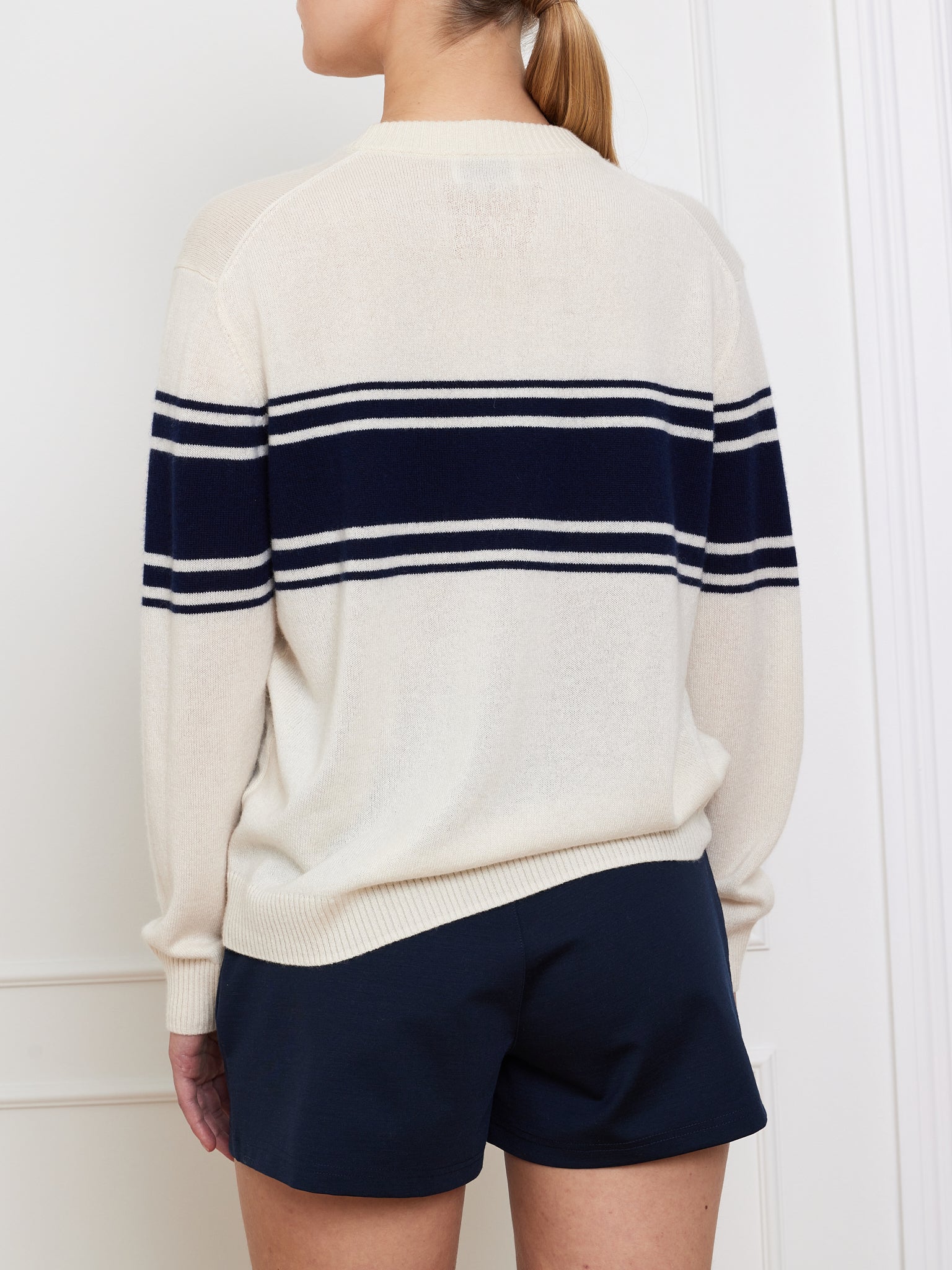 A person with long hair tied back faces away from the camera, wearing the We Norwegians' Yacht Sweater Women, a cream-colored cotton blend with black stripes, paired with dark shorts. A white wall background enhances the simplicity of this classic look.