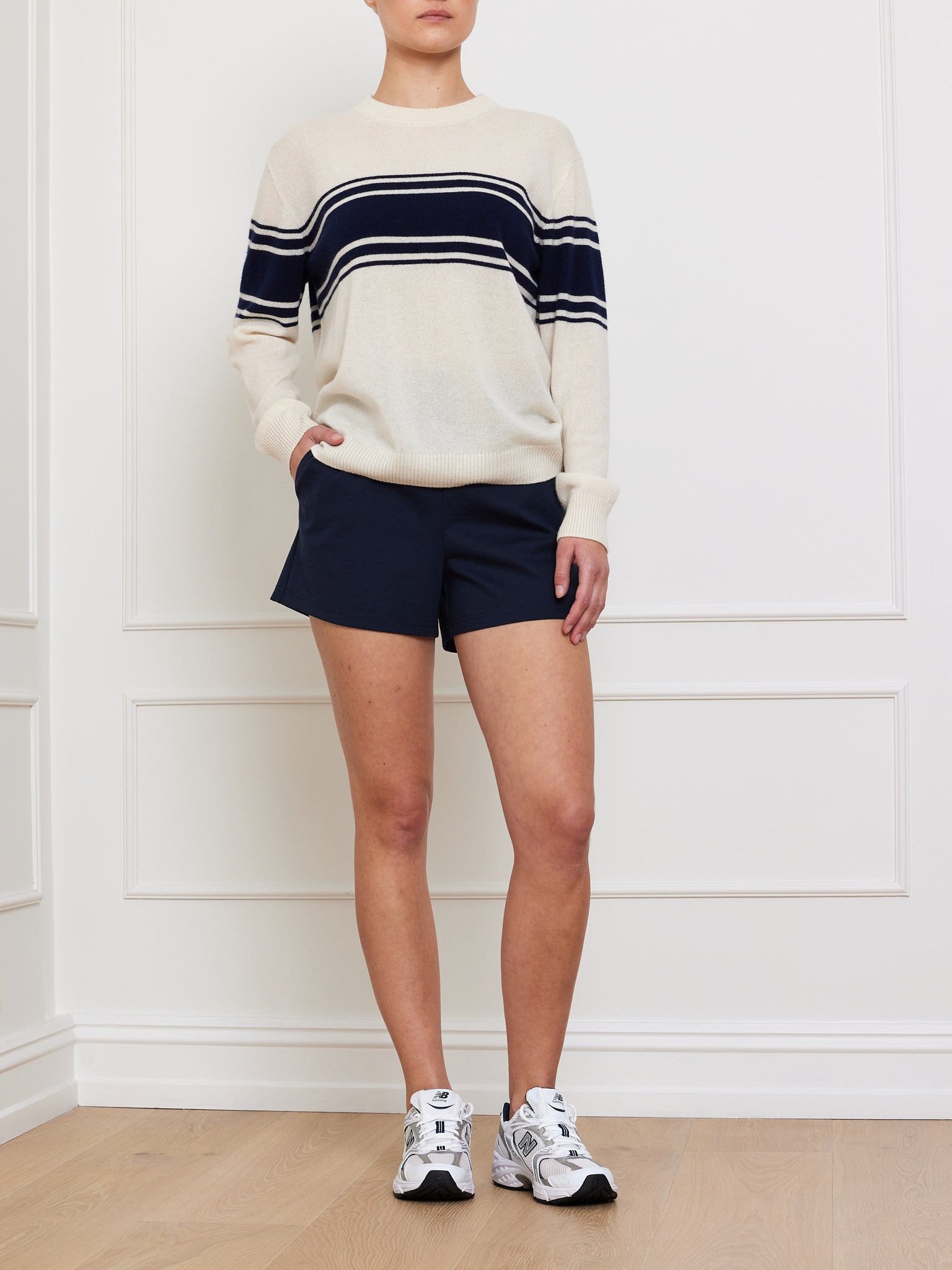 A person stands against a white wall, wearing We Norwegians' Yacht Sweater Women in cream with navy stripes. They pair it with navy shorts and white athletic shoes. One hand is in their pocket, and the other rests by their side on the wooden floor.