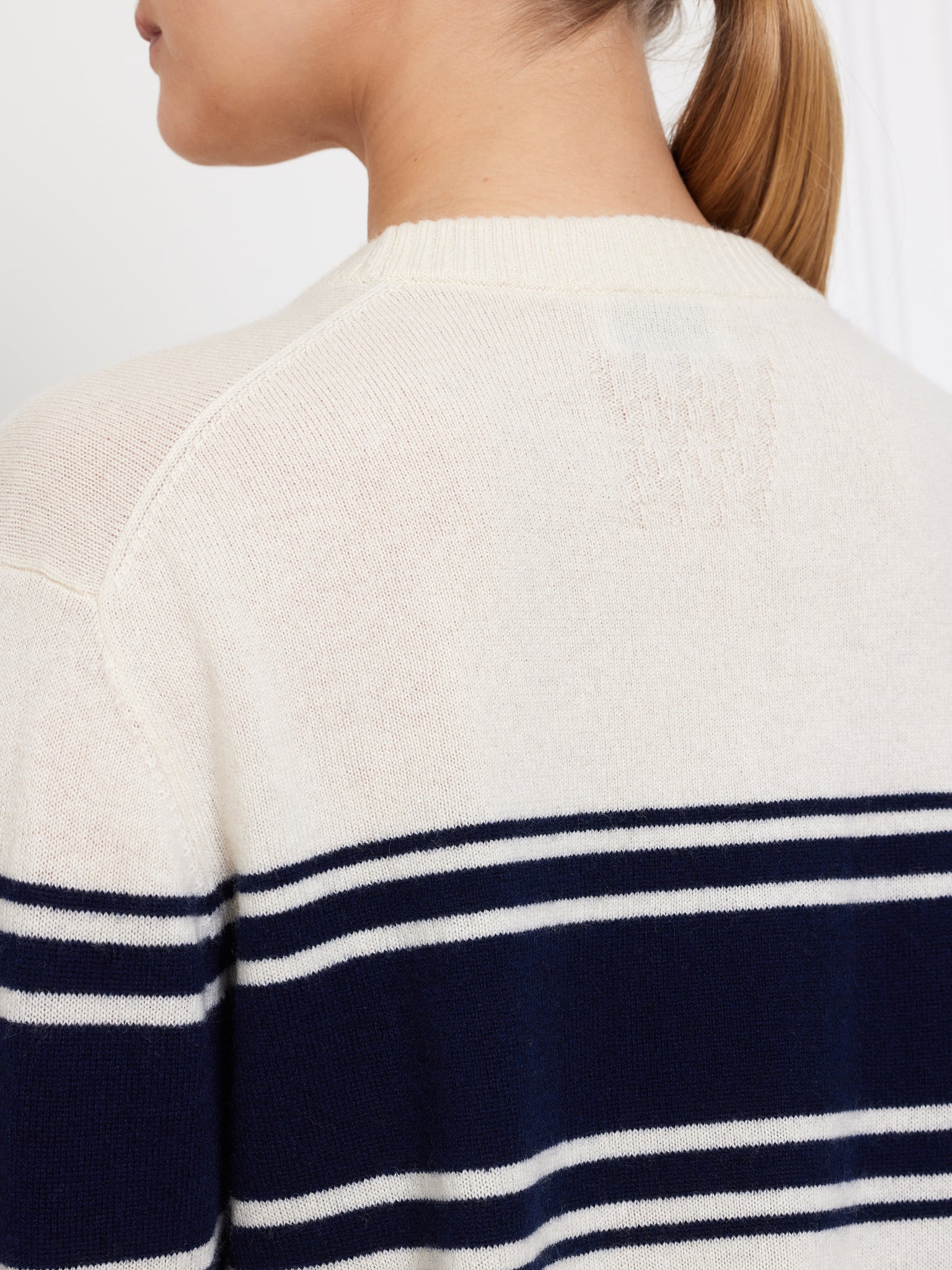 Wearing We Norwegians' Yacht Sweater Women, a light beige merino wool piece with navy and white horizontal stripes across the back, this person has their hair in a ponytail and is turned slightly away from the camera.
