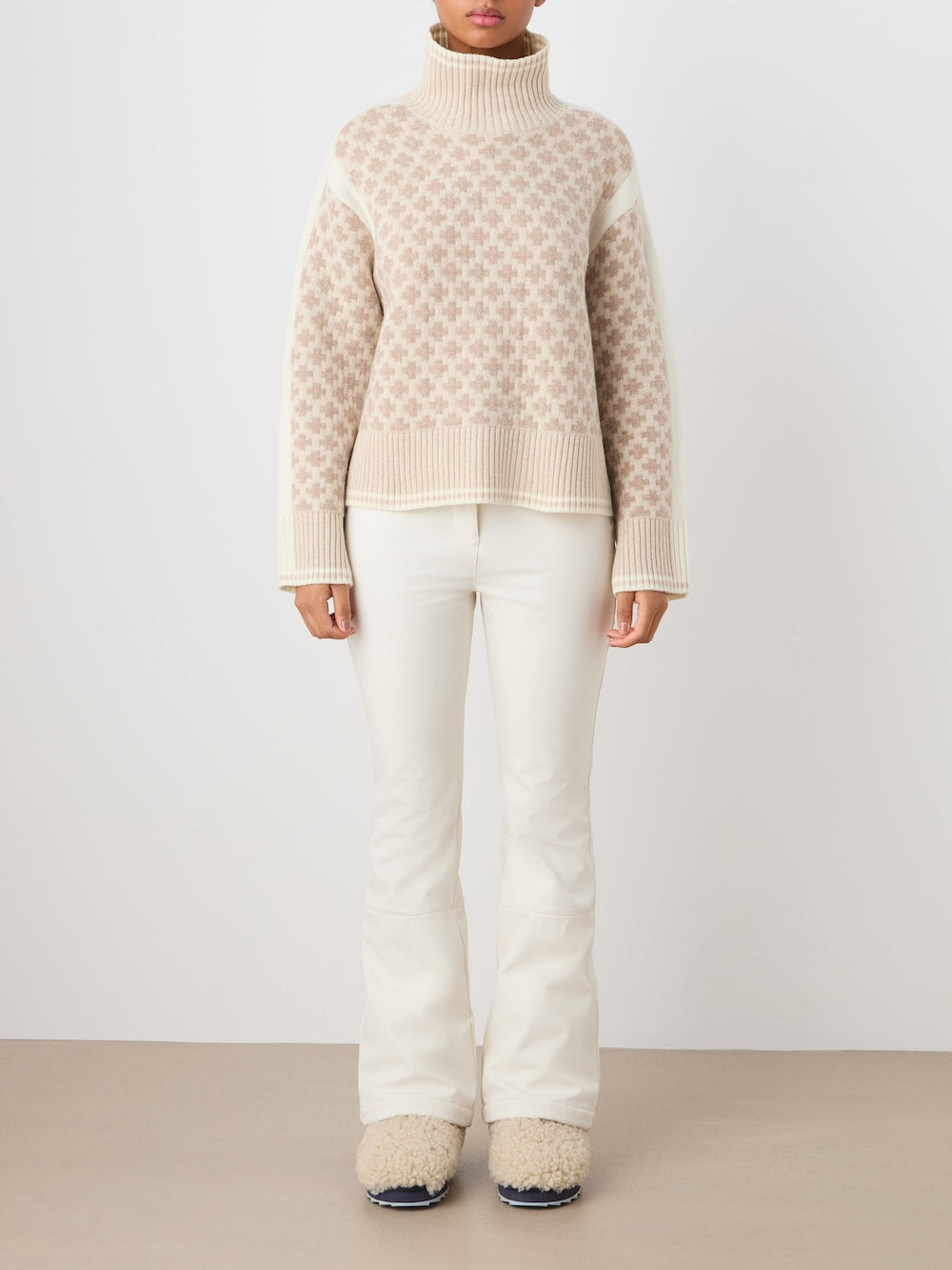 A person stands against a plain background wearing the We Norwegians Boat Sweater women - Pre Spring 26, featuring an oversized beige and white checkered knit turtleneck. They pair it with white flared pants and cream textured shoes. Face not visible.
