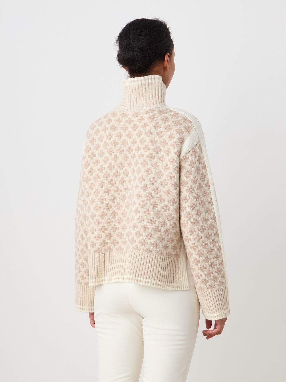 A woman with dark hair in a low bun, seen from behind, wears the We Norwegians Boat Sweater women - Pre Spring 26 in beige and white with white pants against a plain white background.
