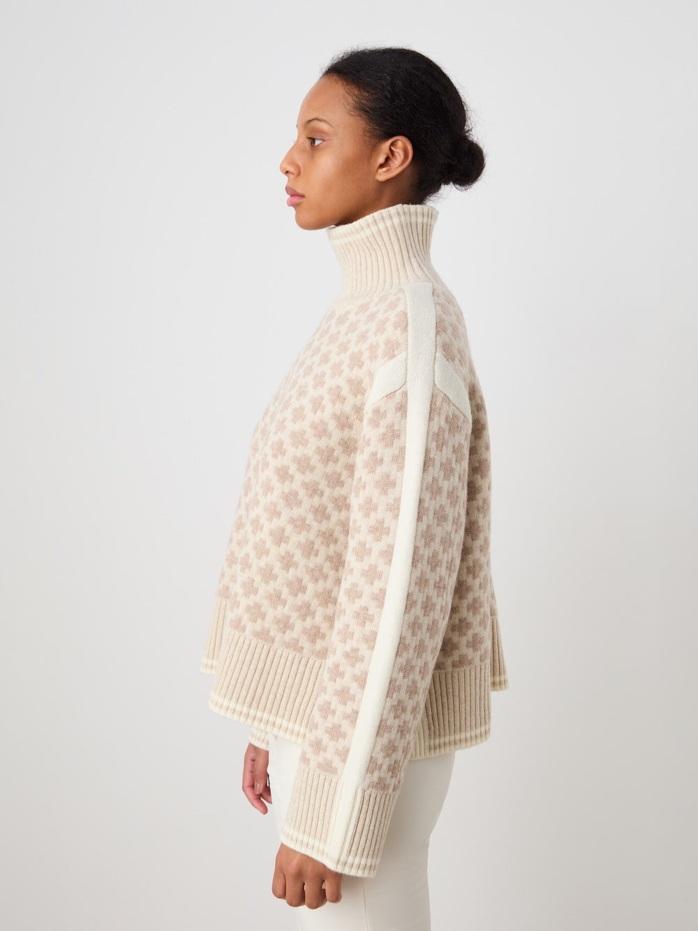 A woman in profile wears the We Norwegians Boat Sweater women - Pre Spring 26, an oversized merino cashmere knit with a high neck, beige and white pattern, ribbed cuffs and hem, and a vertical white sleeve stripe against a plain light background.