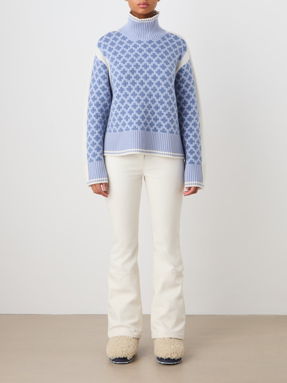 A person wears the We Norwegians Boat Sweater women - Pre Spring 26, an oversized blue and white patterned turtleneck, paired with cream flared pants and fuzzy slippers. Their head is not visible; they stand against a plain background.