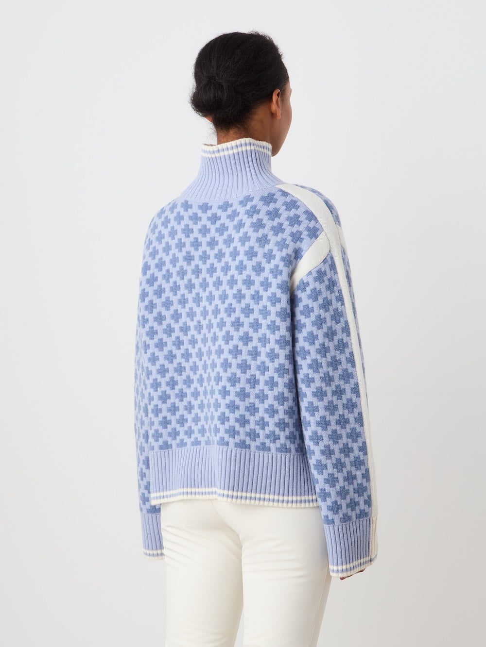 A person with dark hair in a low bun wears the We Norwegians Boat Sweater women - Pre Spring 26, a blue and white patterned oversized knit turtleneck, with white pants, facing away from the camera against a plain white background.