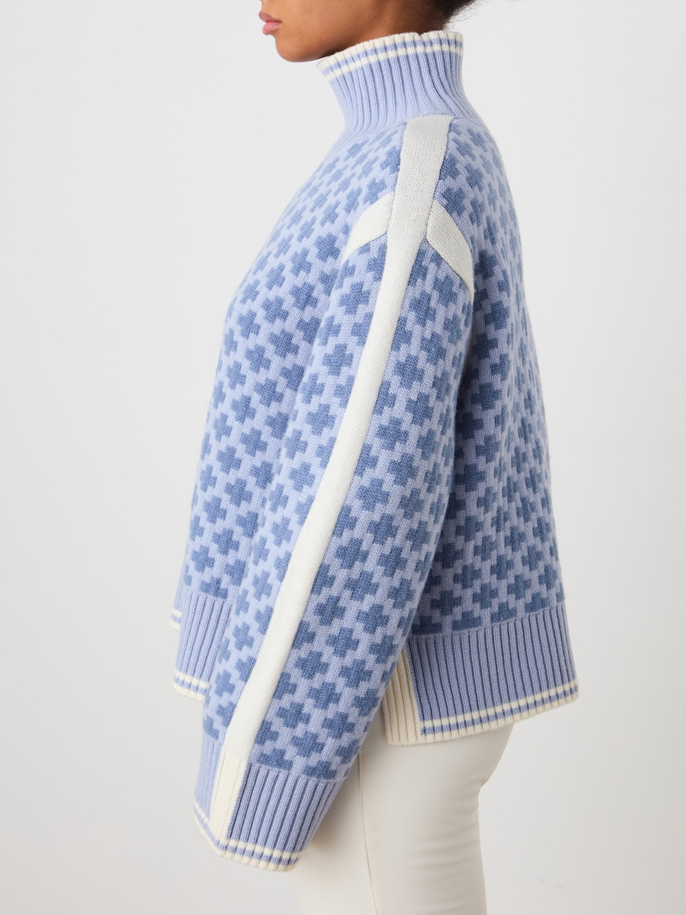 A woman stands sideways against a white background, wearing the We Norwegians Boat Sweater women - Pre Spring 26, an oversized blue and white houndstooth-patterned turtleneck with white stripes on the shoulders and sleeves.