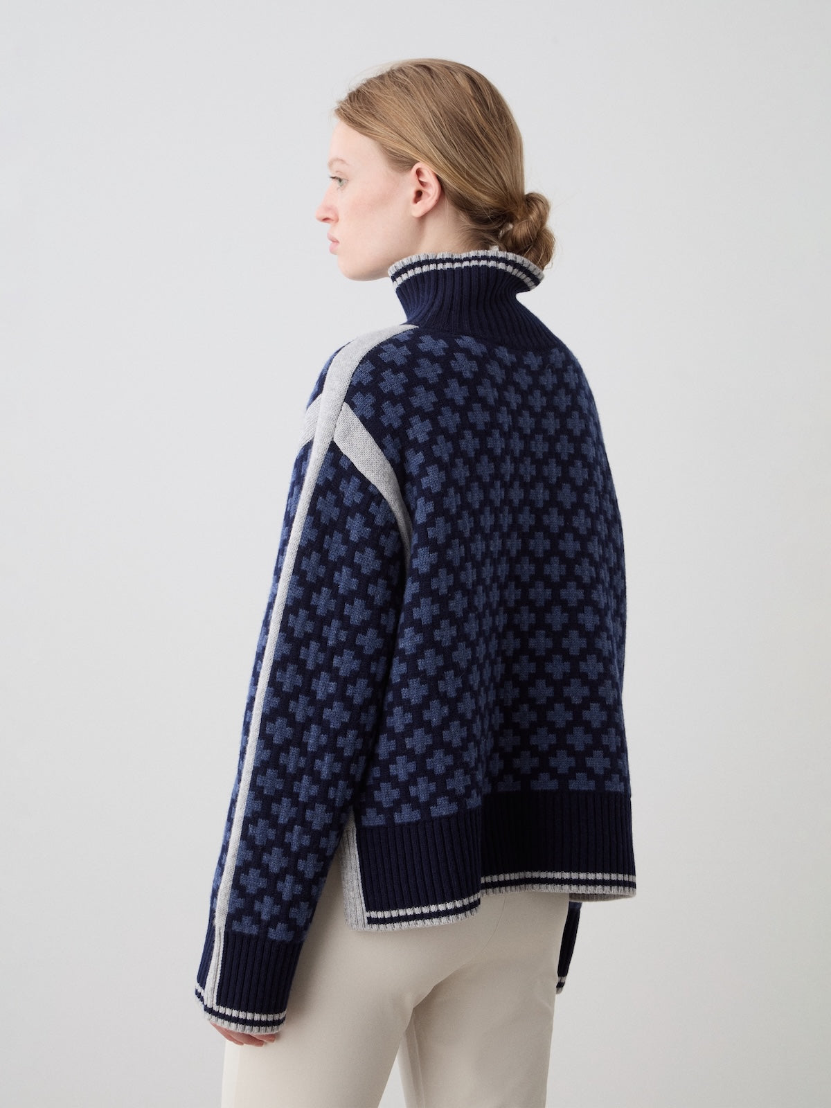 A person with light brown hair in a low bun faces away, wearing the We Norwegians Boat Sweater Women—an oversized navy blue knit with a geometric cross pattern and white striped sleeves—paired with light-colored pants.