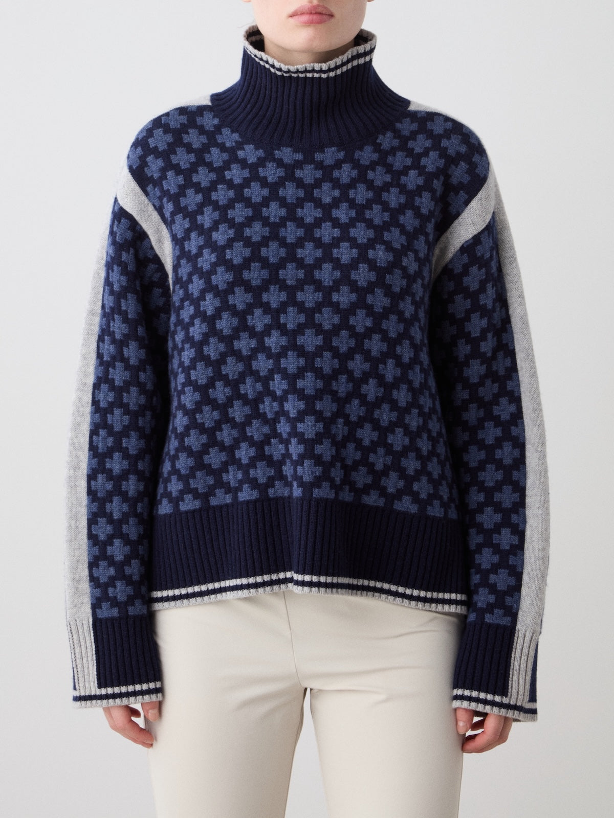 A woman wears the We Norwegians Boat Sweater Women—navy blue merino cashmere with a light blue geometric cross, gray sleeves, and white trim—paired with cream pants against a plain background.