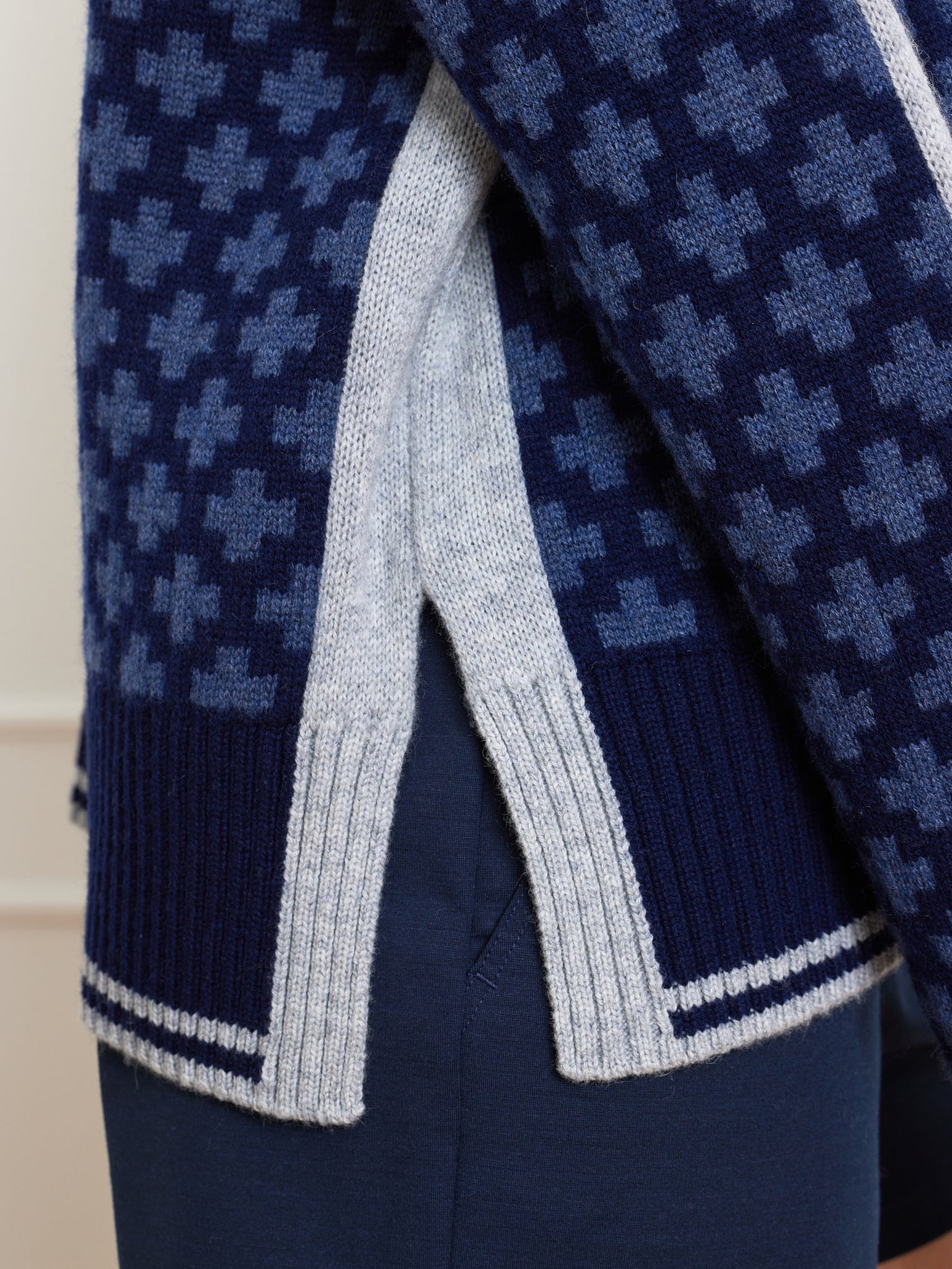 A person wears the We Norwegians Boat Sweater Women, featuring a dark blue merino/cashmere blend with a light blue cross pattern. Reminiscent of a Fisherman's sweater, it sports a ribbed hem and gray side slits, stylishly paired with dark pants.