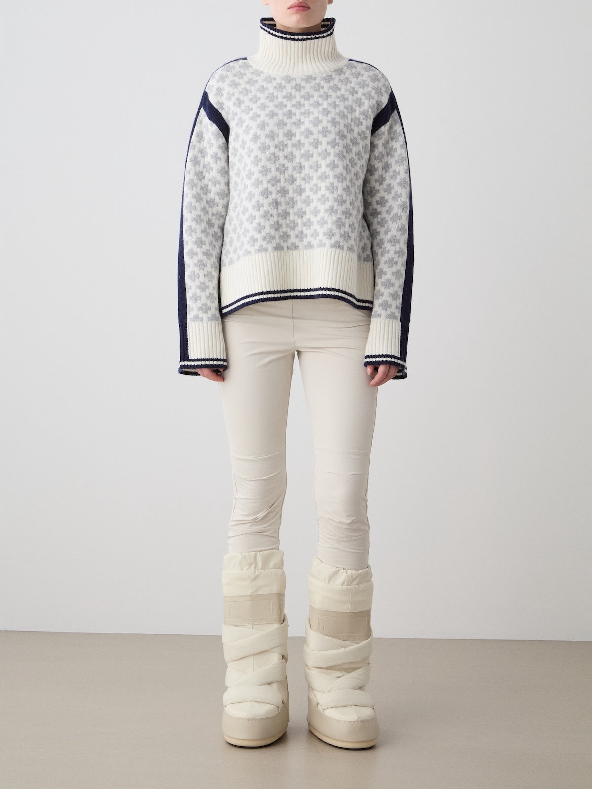 A person stands against a plain background wearing the We Norwegians Boat Sweater Women in gray and white, paired with cream pants and tall padded cream winter boots. Their face is partially out of the frame.