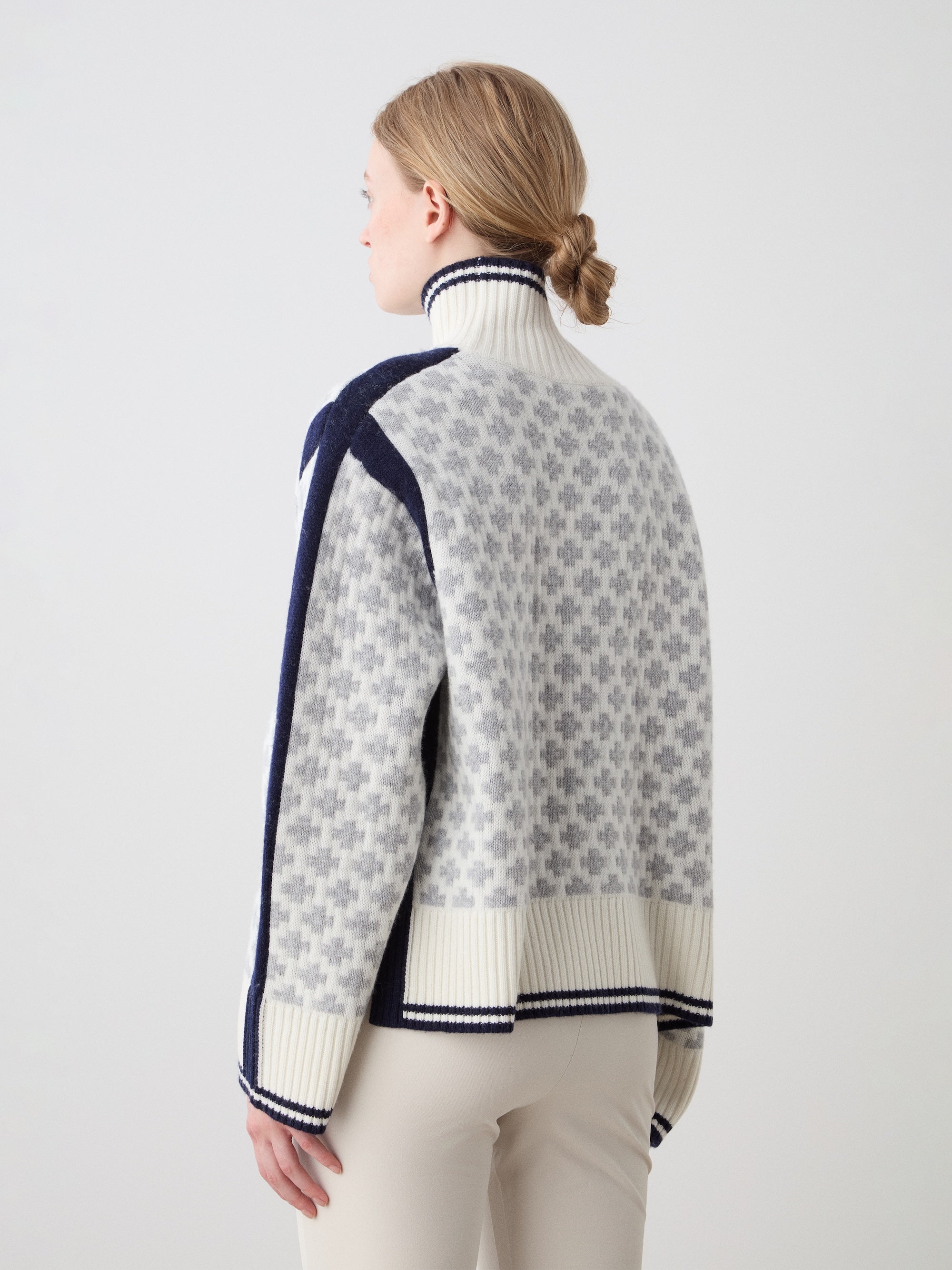 A person with light hair in a low bun is seen from the back wearing the We Norwegians Boat Sweater Women, a light gray and navy merino cashmere sweater with vertical geometric patterns. The background is plain white.