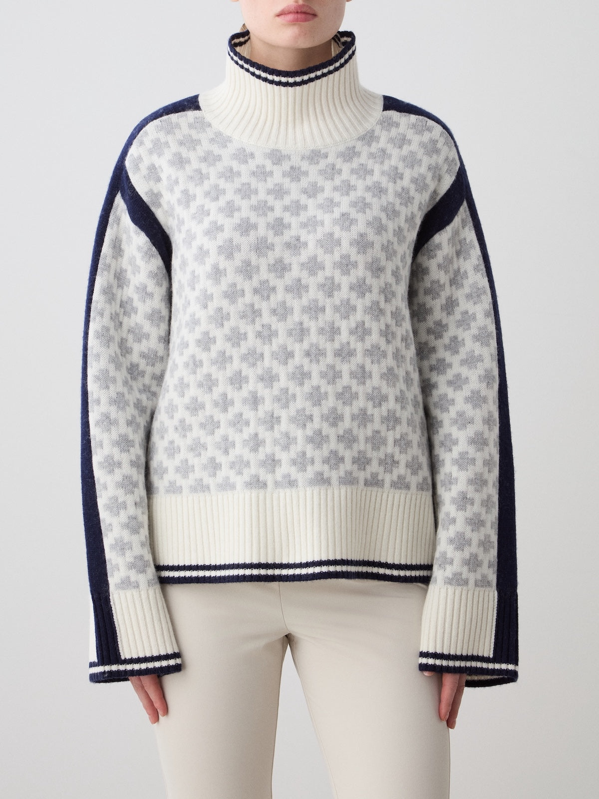 A person wearing the We Norwegians Boat Sweater Women—an oversized knit with a white and gray geometric pattern, navy blue sleeve accents, and cream ribbed cuffs and hem—paired with light-colored pants.