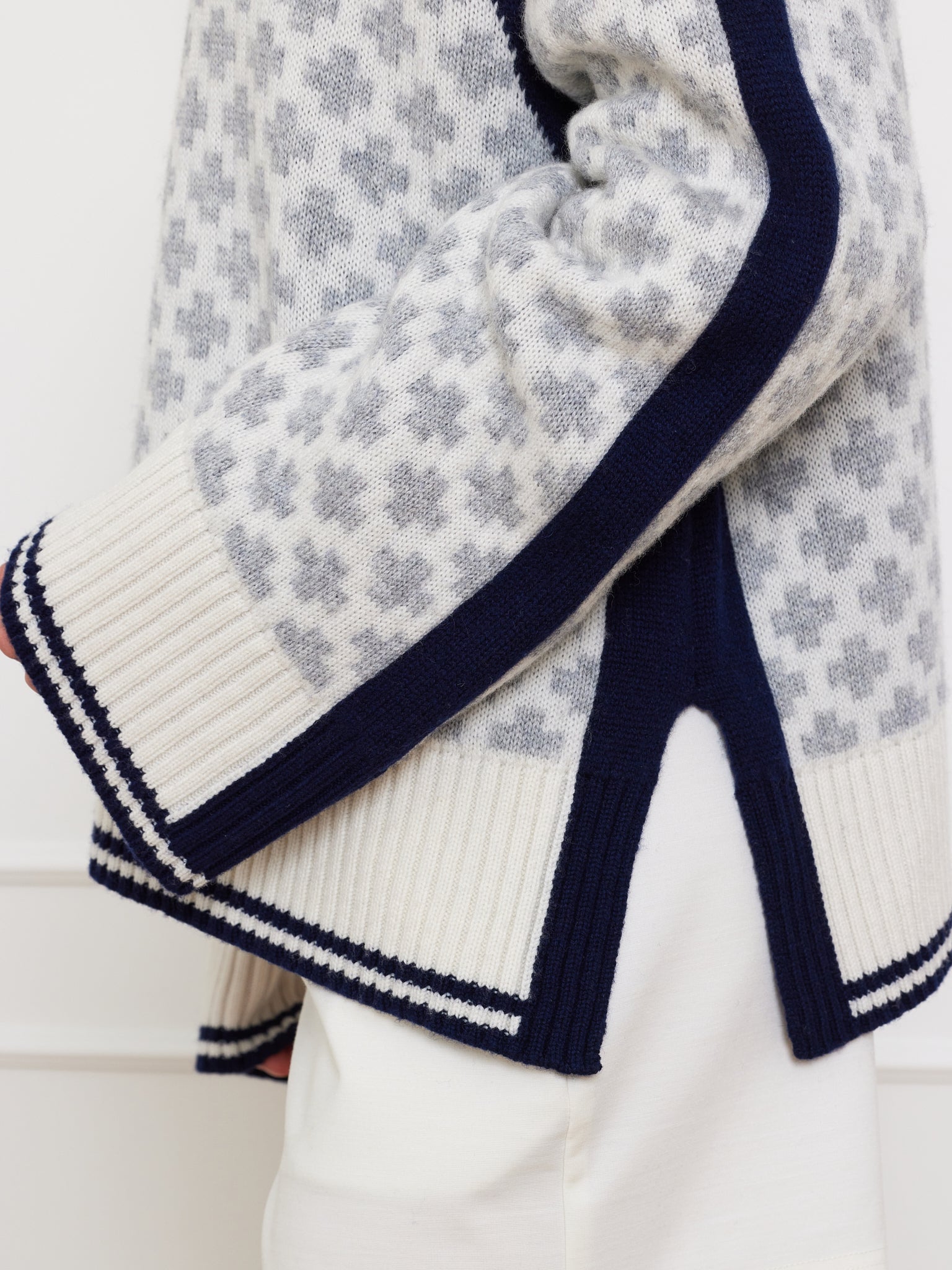 A close-up shows a person wearing the We Norwegians Boat Sweater Women in light gray, made from luxurious merino/cashmere yarn. It features dark blue and white striped accents on the cuffs and hem, a side slit, and pairs elegantly with white pants for an ideal Scandi Summer look.