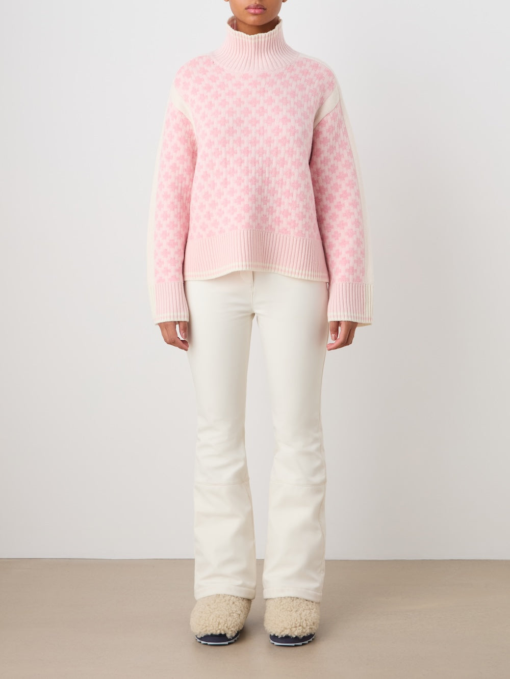 A person wears the We Norwegians Boat Sweater women - Pre Spring 26, featuring pink and white checks, with cream flared pants and beige fuzzy slippers against a white background. The person's head is not visible.