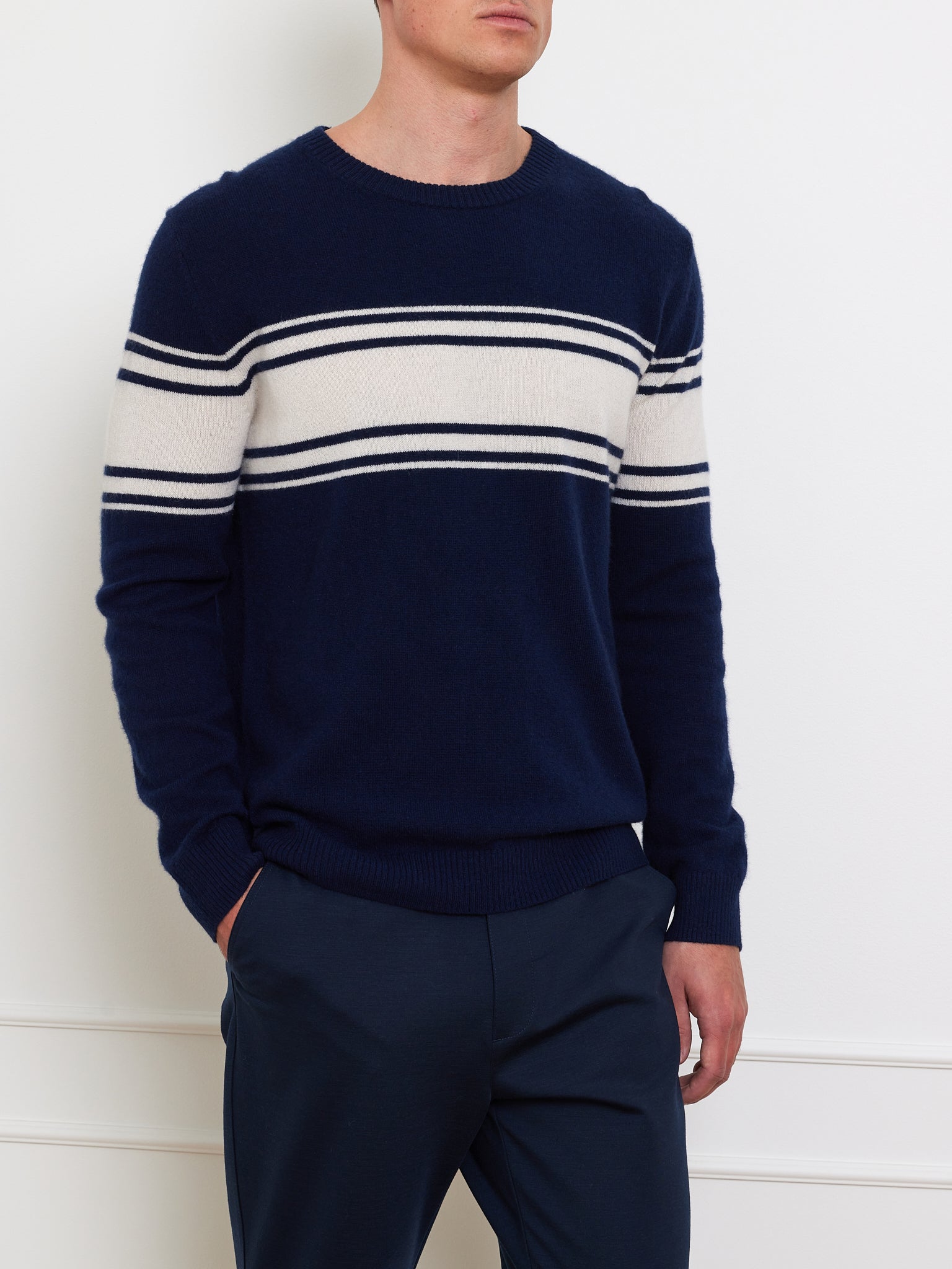 A person wears the We Norwegians Yacht Sweater Men, featuring classic navy blue with white and navy stripes on the chest and arms. With a hand in their dark pants pocket, they stand against a light wall, highlighting this timeless piece.