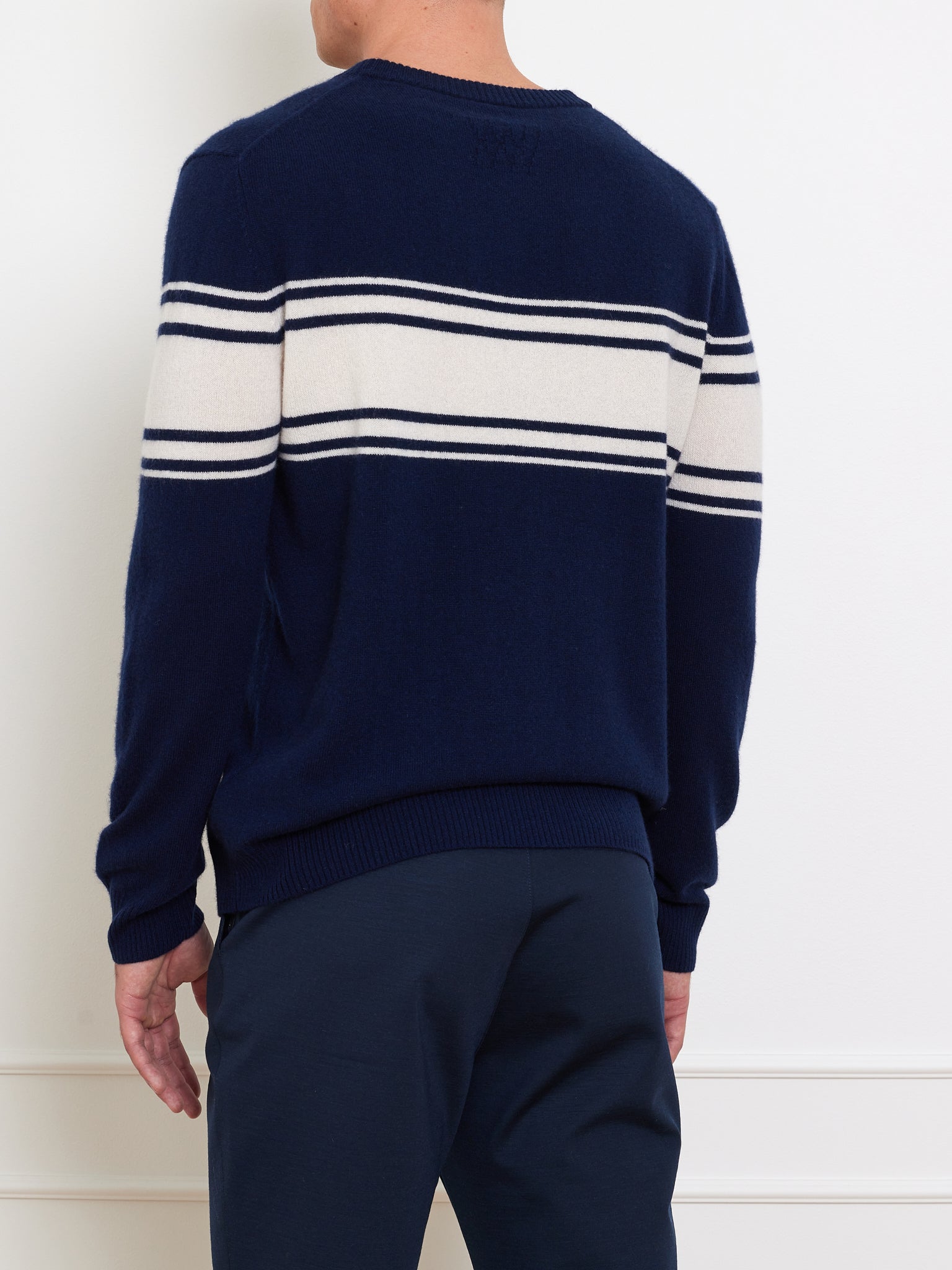 A person wears the Yacht Sweater Men by We Norwegians, a navy blue classic with white stripes across the chest and arms, paired with navy pants. The stylish ensemble is a true wardrobe staple as they stand against a plain white background with their back to the camera.