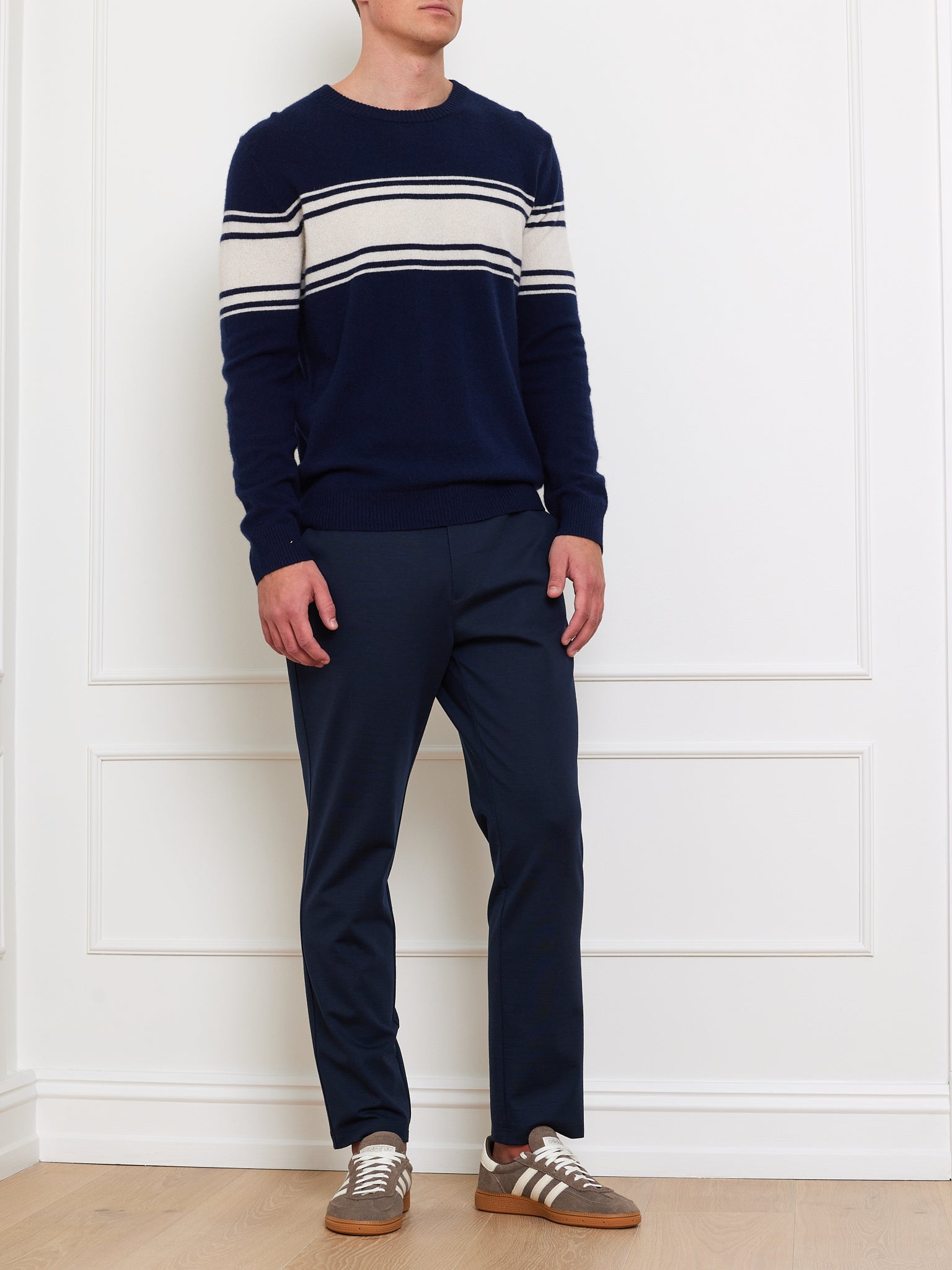 A person wears the We Norwegians Yacht Sweater Men, a classic navy piece with white stripes on the chest and arms—a wardrobe staple—paired with navy pants and brown sneakers, standing against a white paneled wall on light wood flooring.
