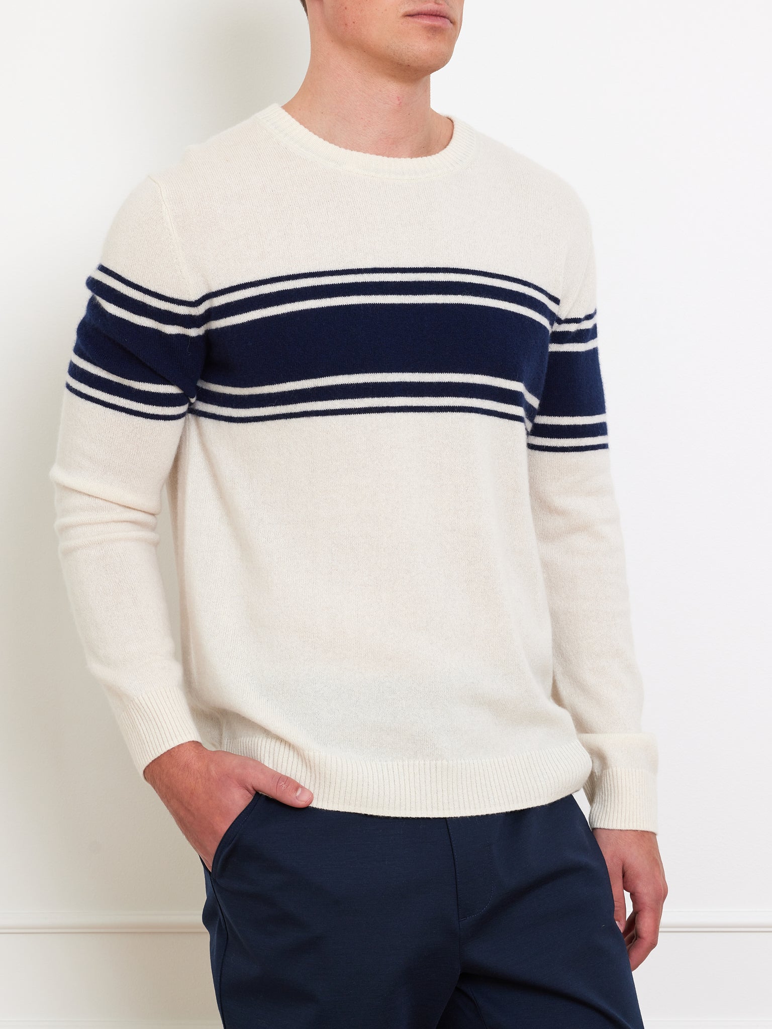Wearing the We Norwegians Yacht Sweater Men in classic cream with navy stripes, a person pairs it effortlessly with dark pants. Their hands rest casually in their pockets against a plain white wall, embodying a timeless wardrobe staple.