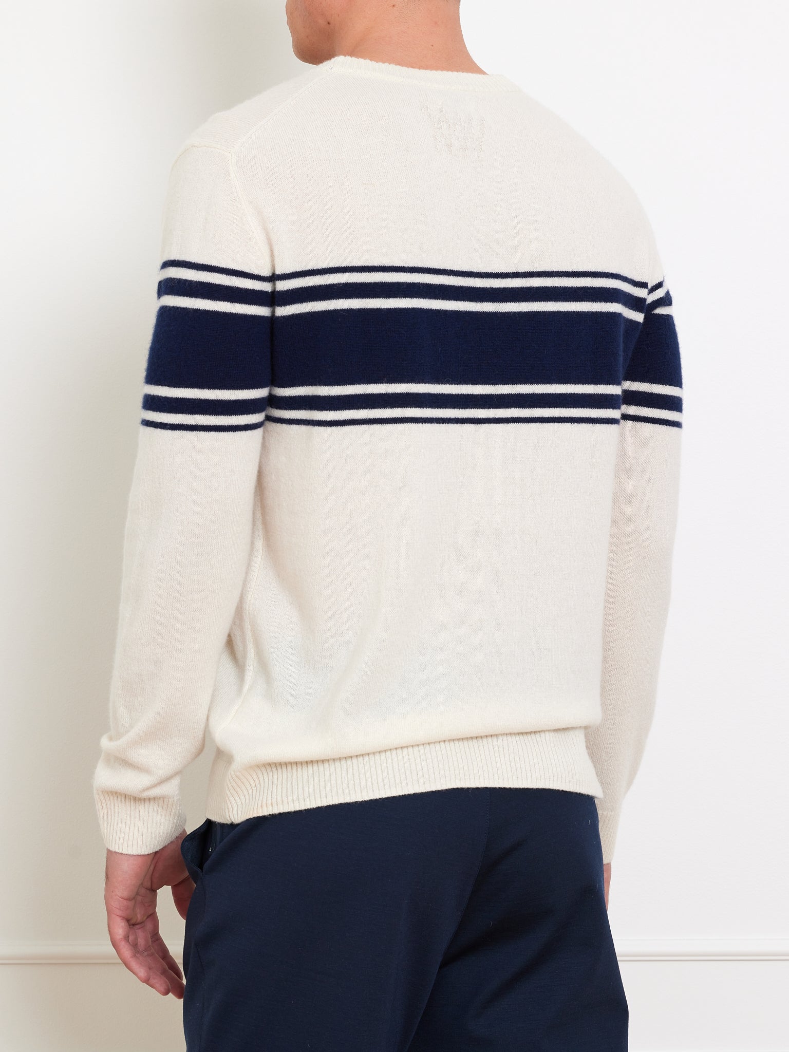 A man wearing the We Norwegians Yacht Sweater Men in classic cream with navy stripes across the back and shoulders pairs it with navy pants against a white background.