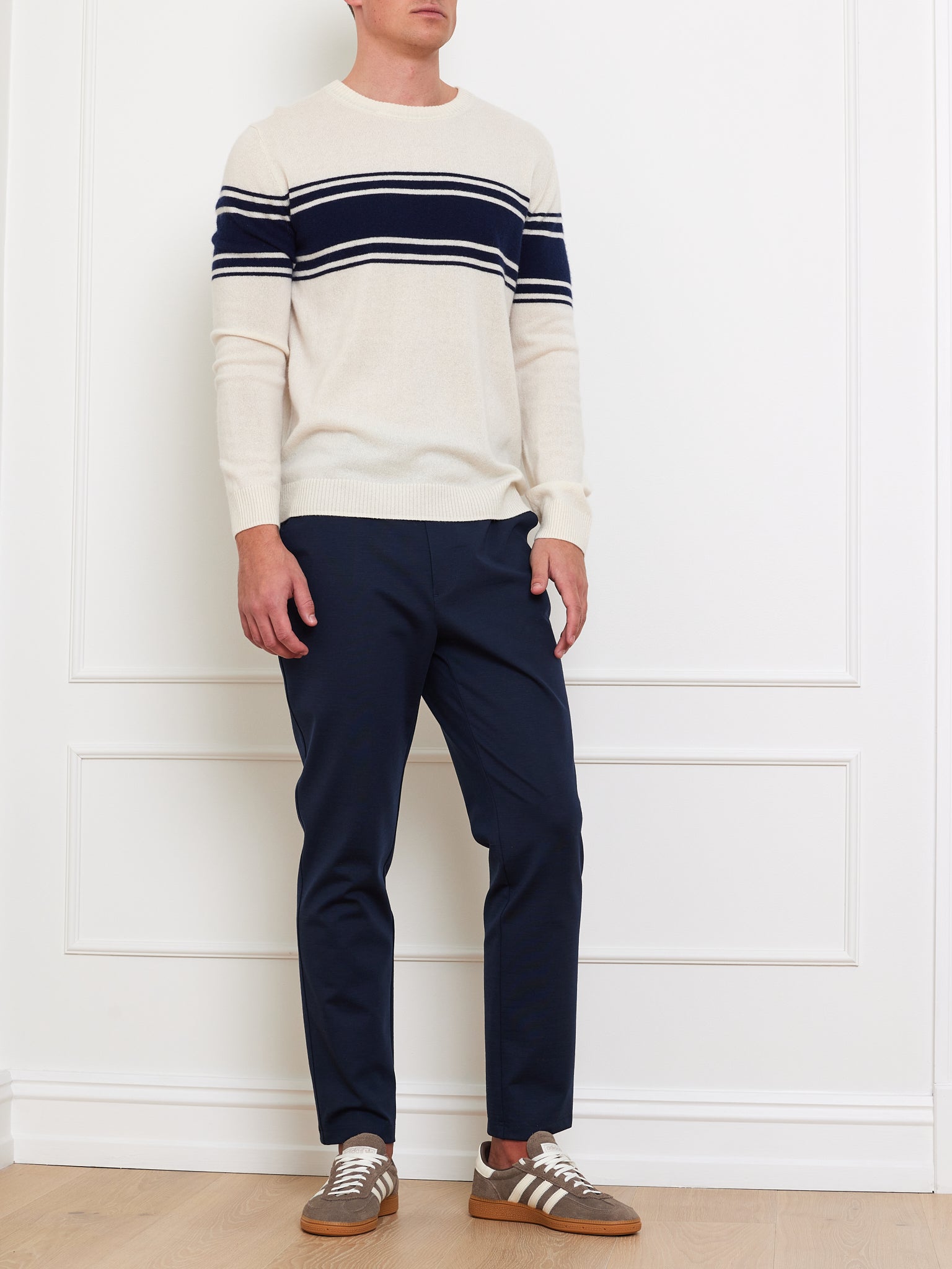 A person wearing a We Norwegians' Yacht Sweater Men—a classic cream sweater with navy stripes—paired with navy trousers and brown and white sneakers stands against a white paneled wall. The person's face is hidden, emphasizing the outfit's timeless appeal.